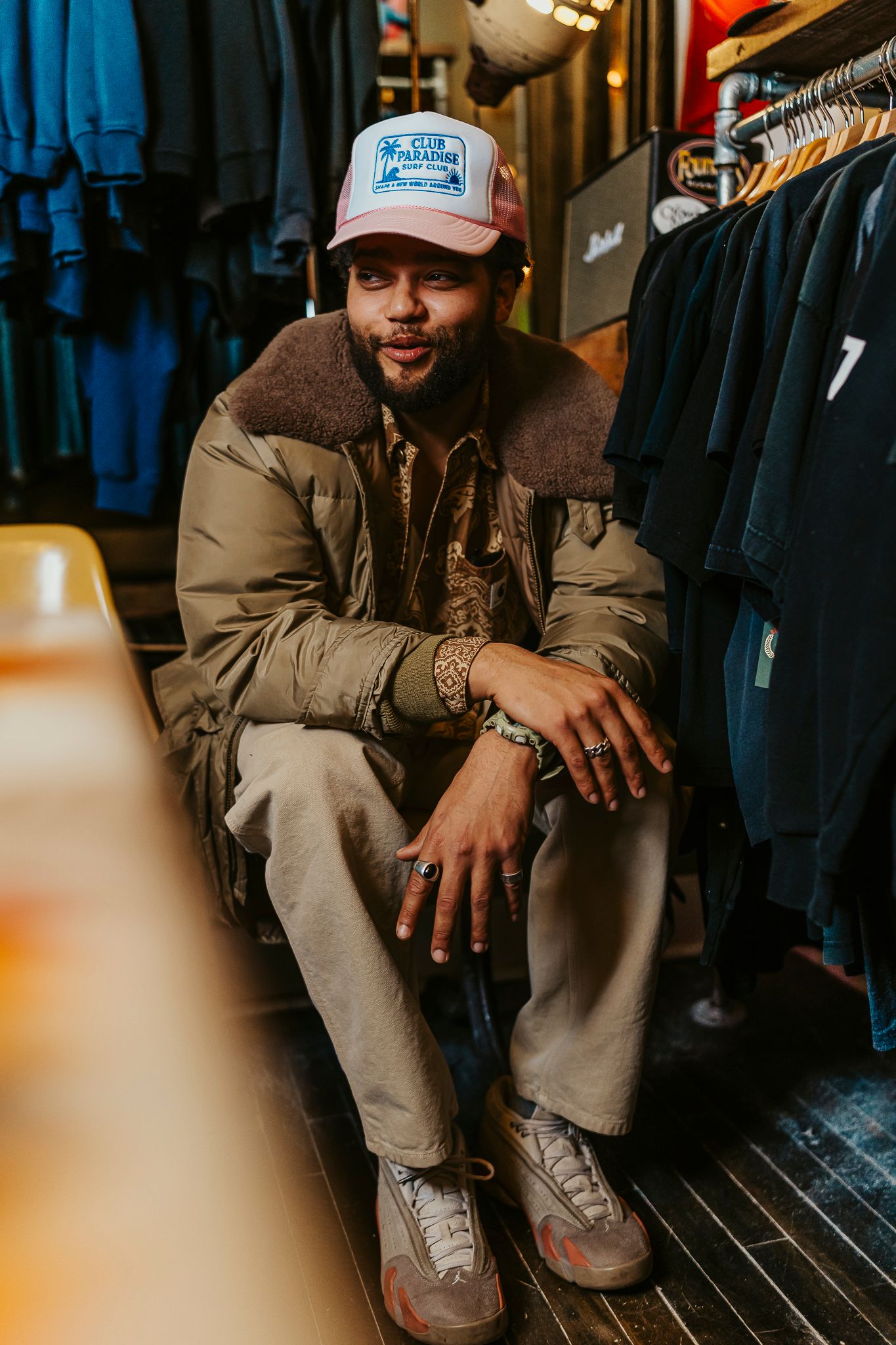 Man in pink trucker hat and olive jacket sits cross-legged in a clothing store, wearing jewelry and sneakers.