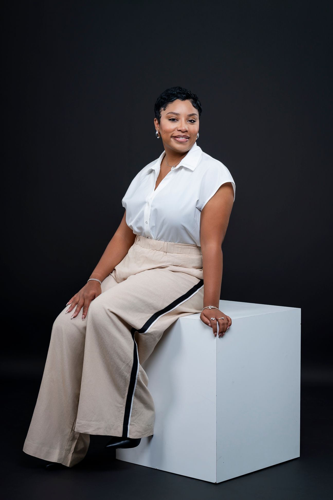A smiling woman in a white blouse and beige wide-leg pants sits on a white cube against a dark background.