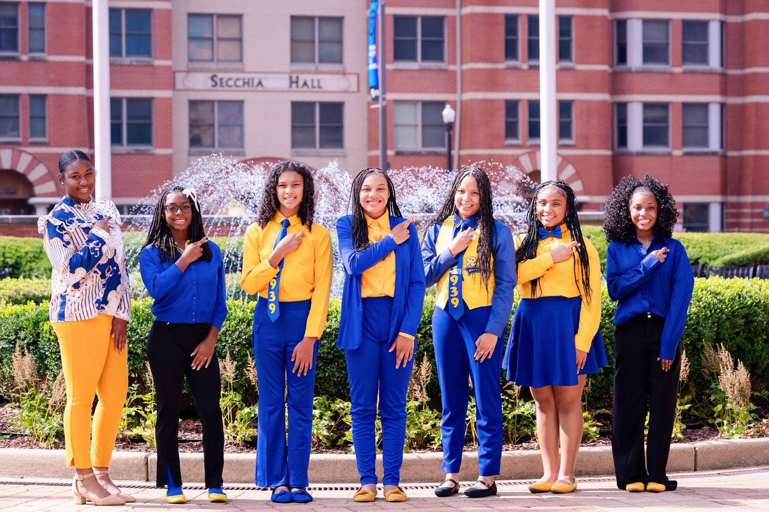 Seven women in matching royal blue and gold outfits pose together outdoors in front of a brick building and fountain on a sunny day.
