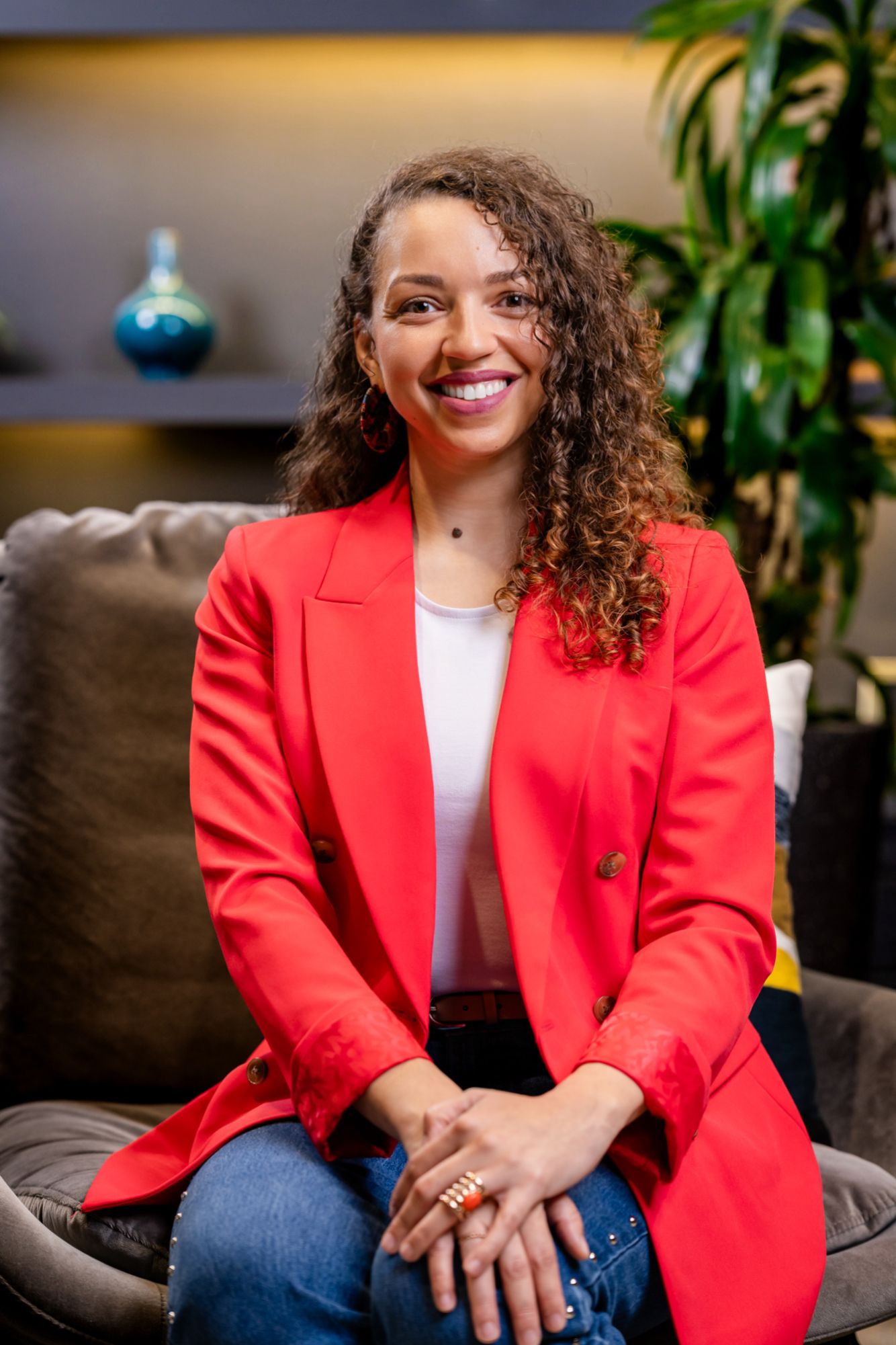 Smiling woman with curly hair in a red blazer and jeans sits on a gray armchair in a modern, warmly lit room with a plant behind her.