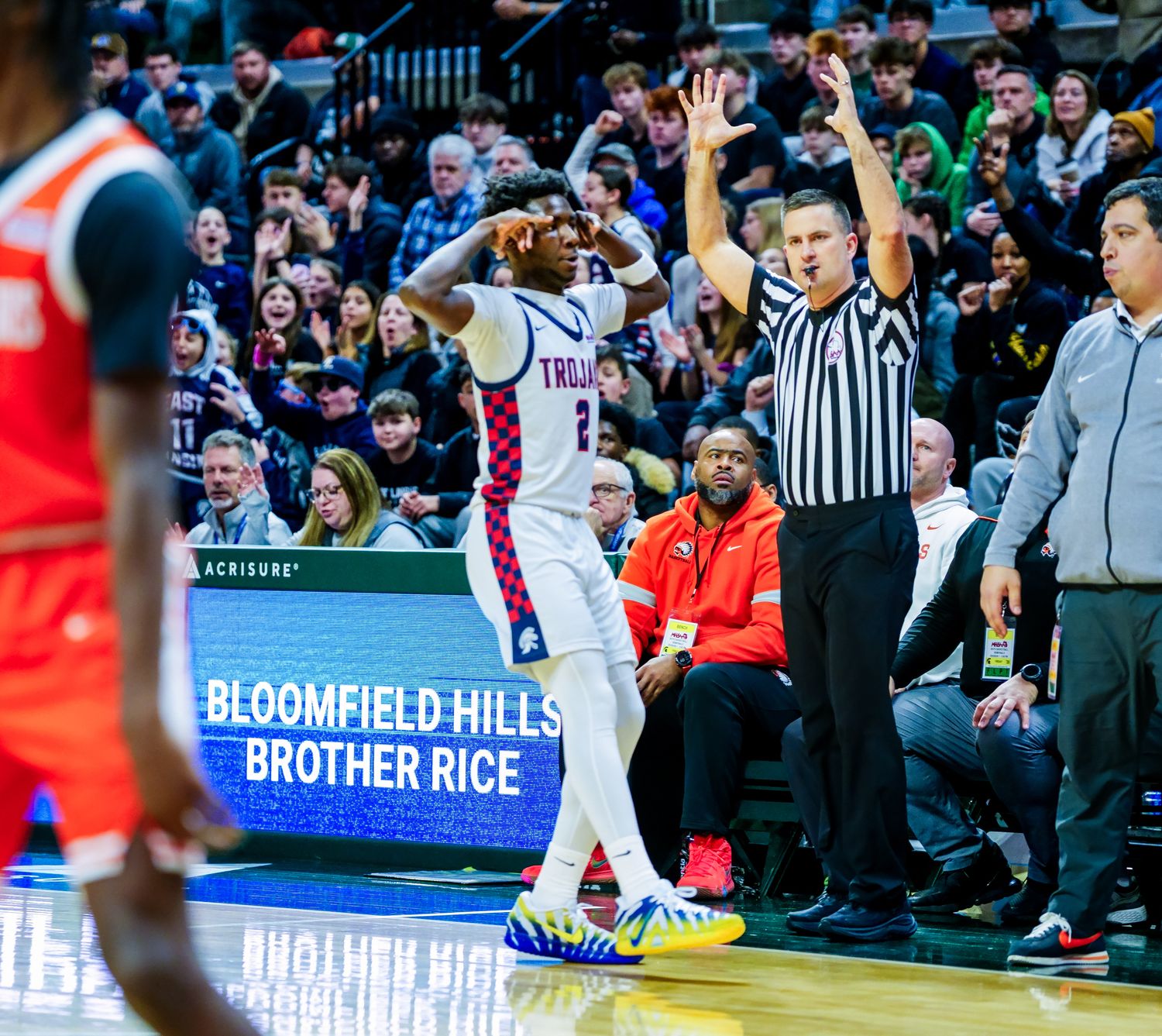 Basketball player celebrates on court near referee, Bloomfield Hills Brother Rice game, crowd cheering in background.