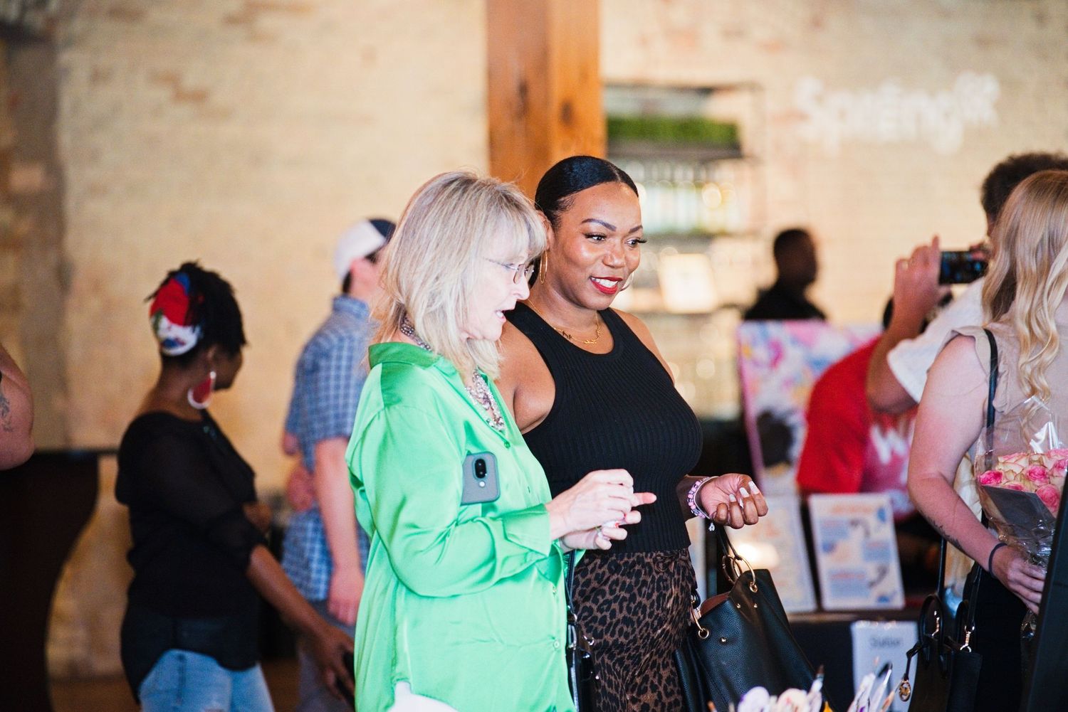 Two women smile and chat at an indoor networking event, one in green and one in a black outfit.