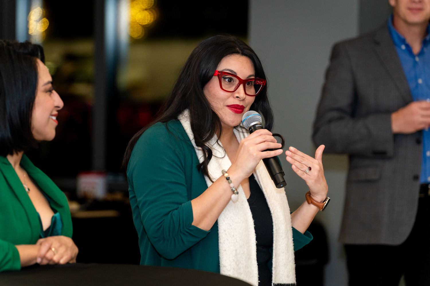 Woman with red glasses speaking into a microphone at an indoor panel discussion event.