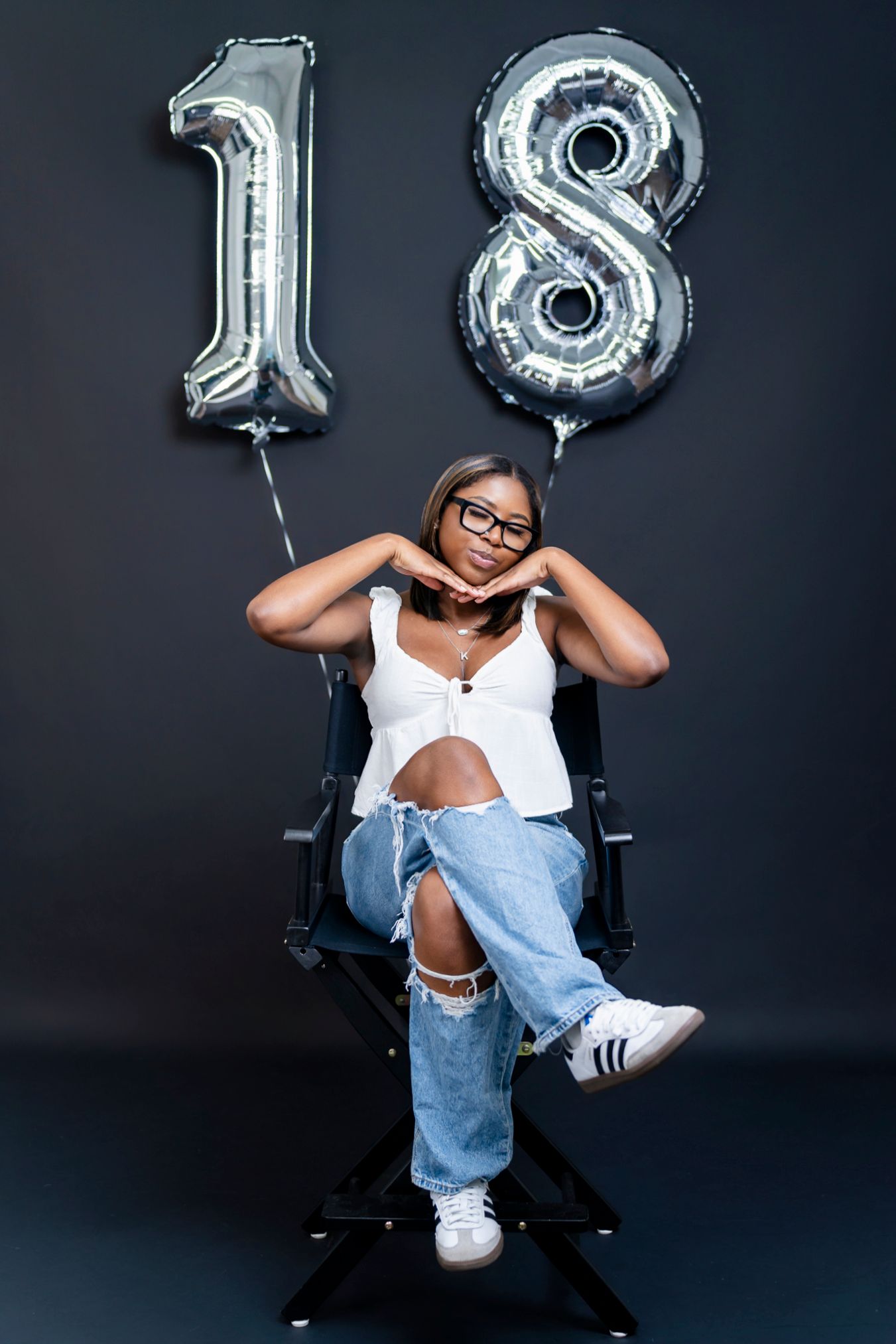Young woman celebrating 18th birthday sits in director chair with large silver number balloons.