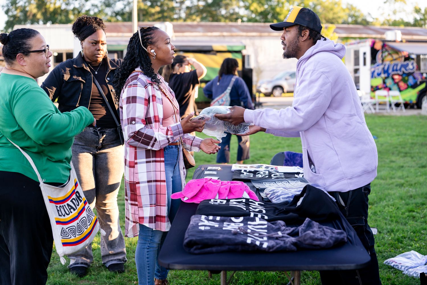 A vendor in a gray hoodie and cap hands an item to a woman in a plaid jacket at an outdoor table of printed apparel while shoppers look on.