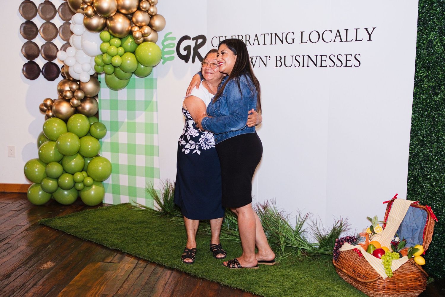 Two women embrace and pose together in front of a sign celebrating locally grown businesses with balloon decor.
