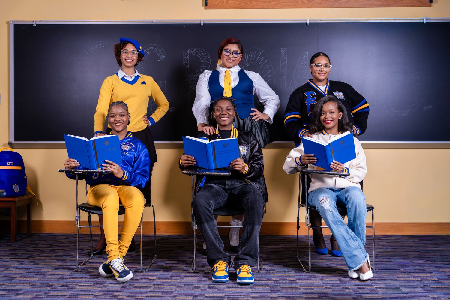 Six students pose in a classroom with blue books, wearing blue and gold outfits in front of a chalkboard.