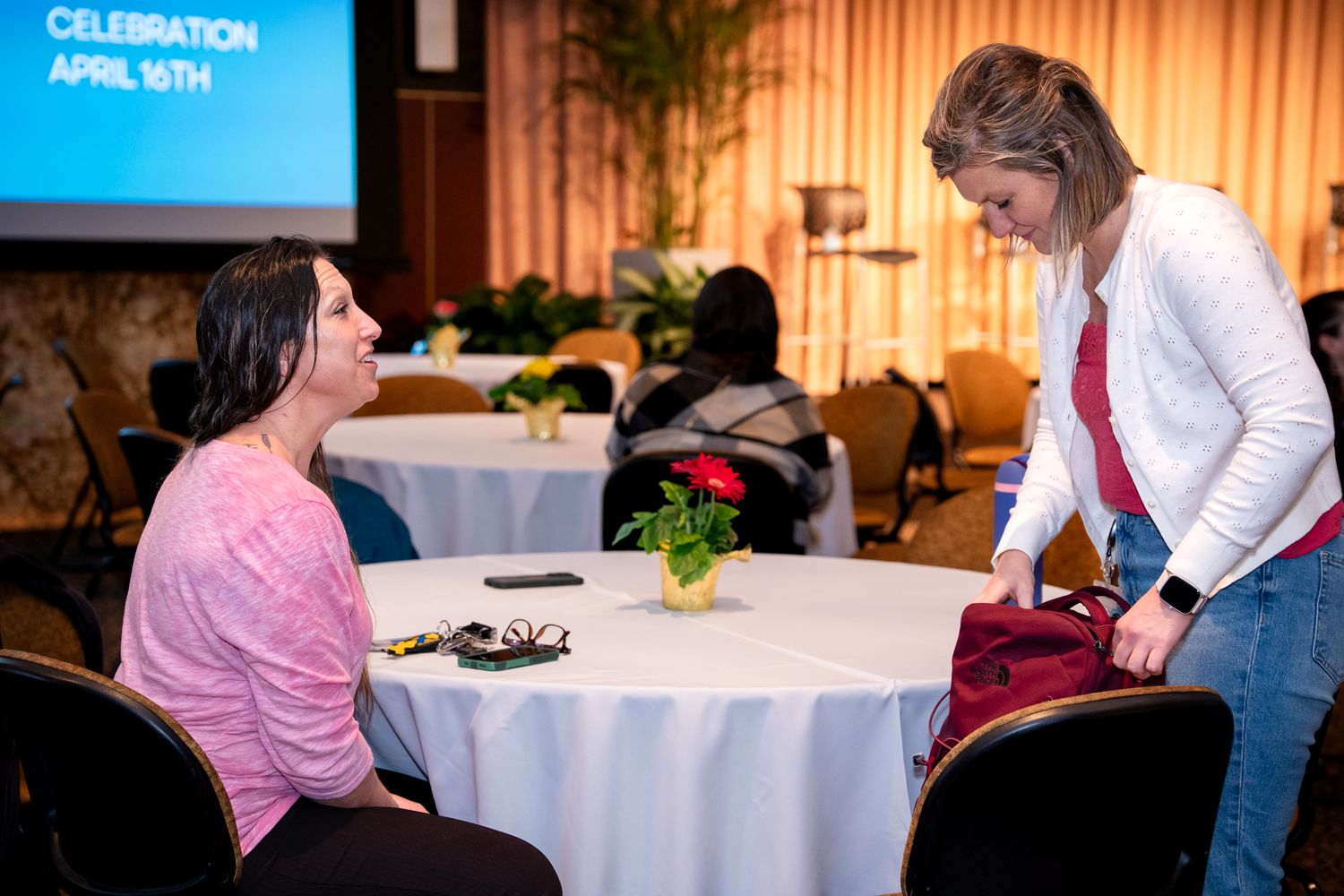 Two women converse at a round table during an April celebration event indoors.
