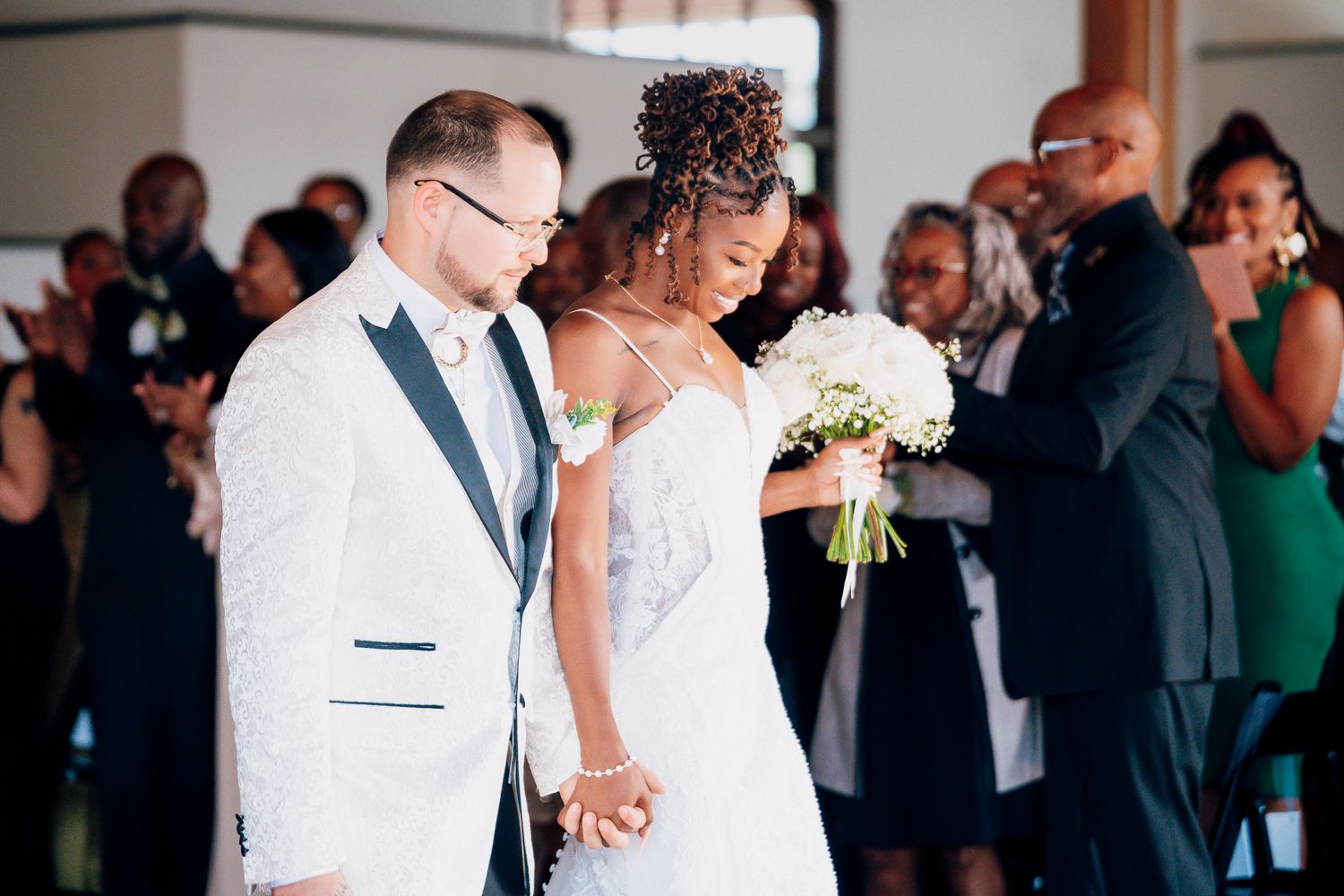 Interracial couple walks hand in hand at wedding ceremony as guests applaud around them.