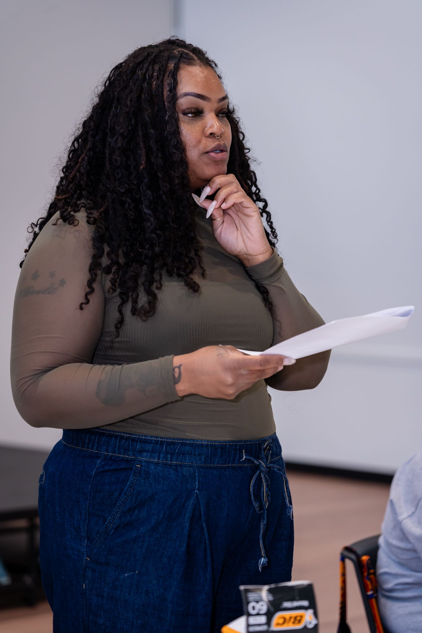 Woman with curly hair holds papers while speaking, wearing olive top and denim pants in a professional setting.
