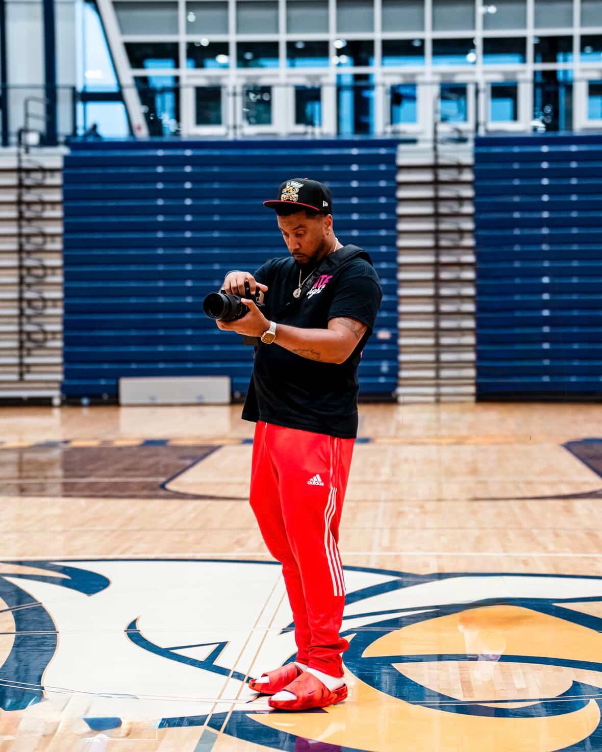 Man in red Adidas pants and black cap reviewing photos on camera while standing on indoor basketball court.