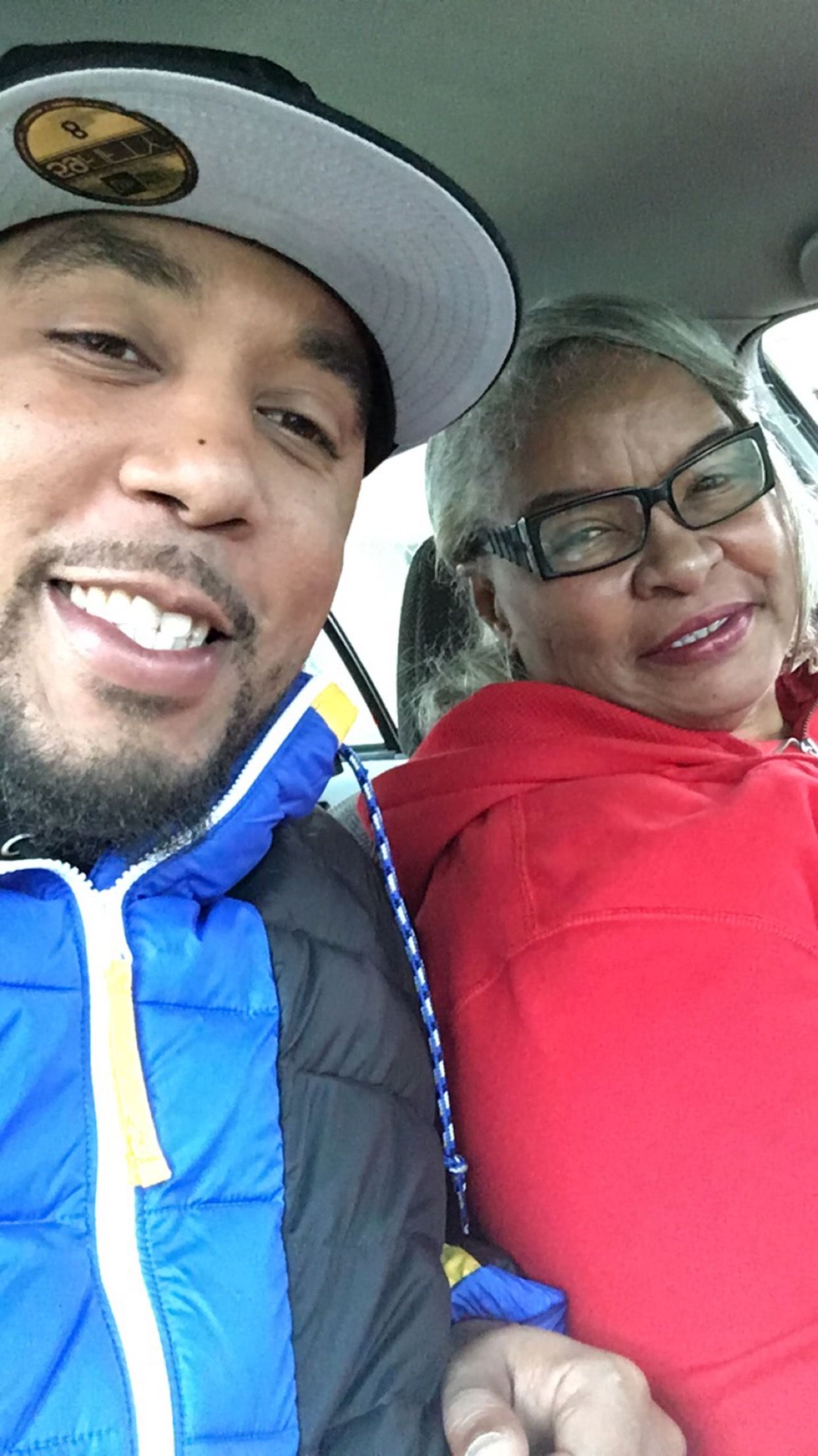A smiling man in a blue jacket and cap takes a selfie with an older woman in a red hoodie wearing glasses inside a car.