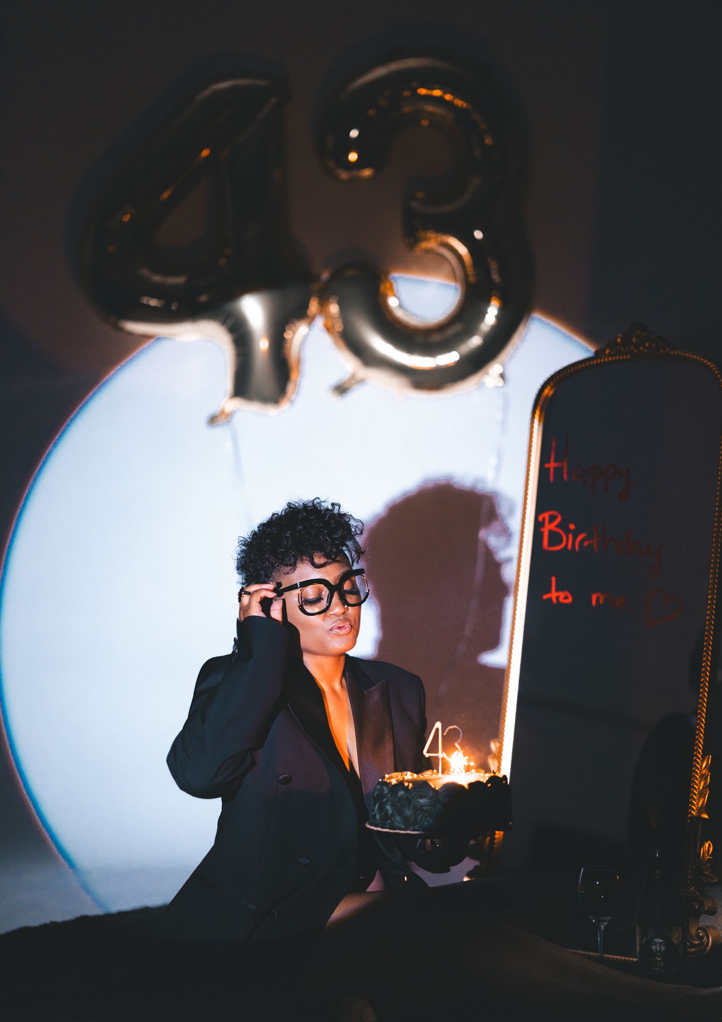 Person in blazer and glasses holds birthday cake near 48 balloons in dark dramatic setting.