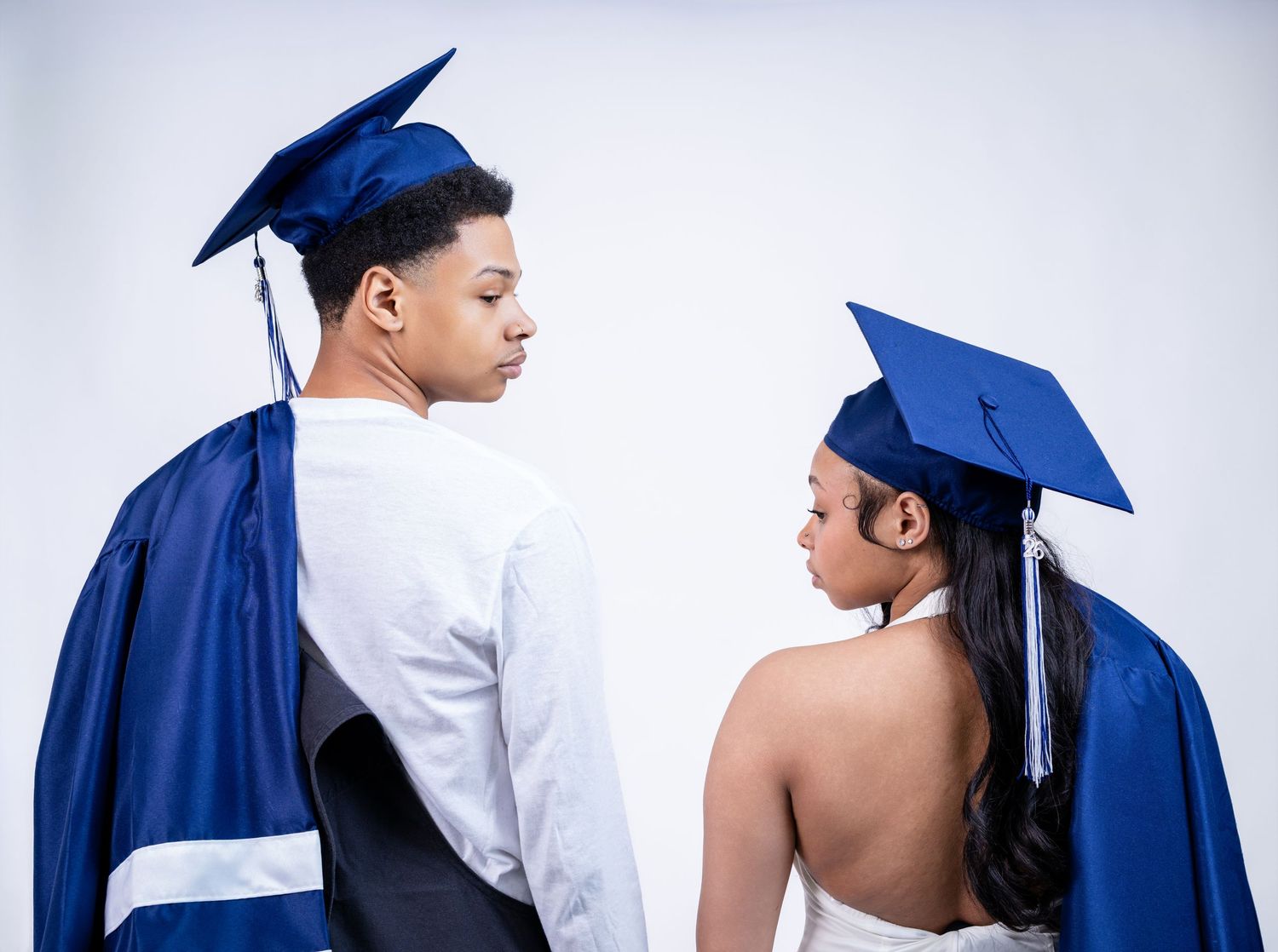 Two graduates in blue cap and gown facing each other against white background.
