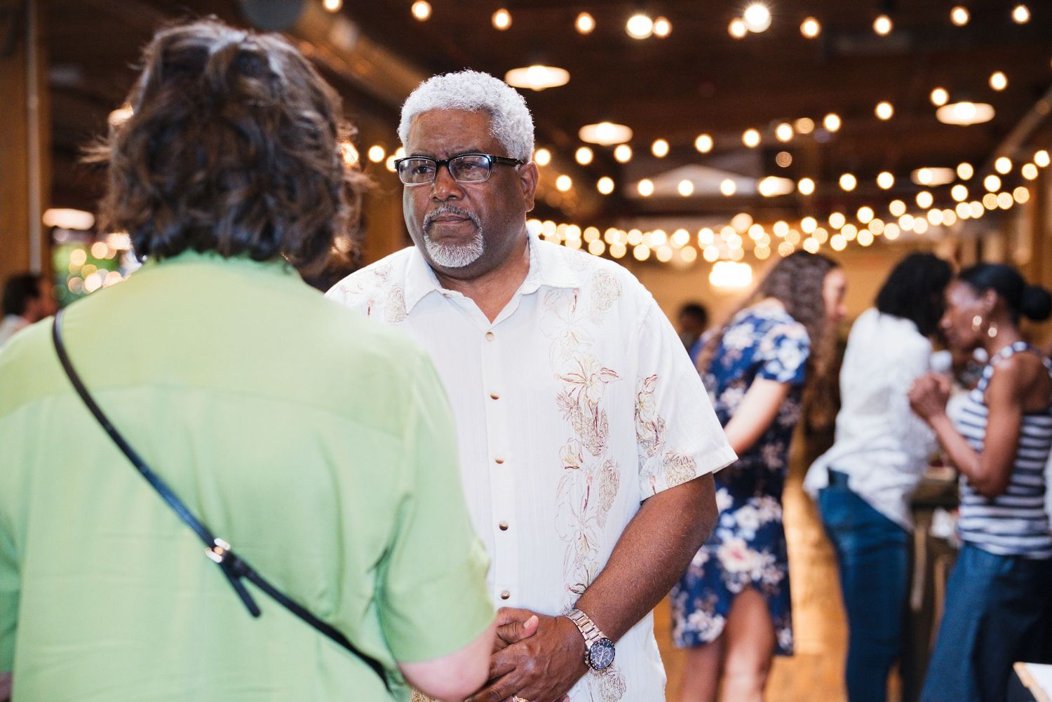 An older man in a white shirt holds hands with a guest at an indoor social gathering with string lights.