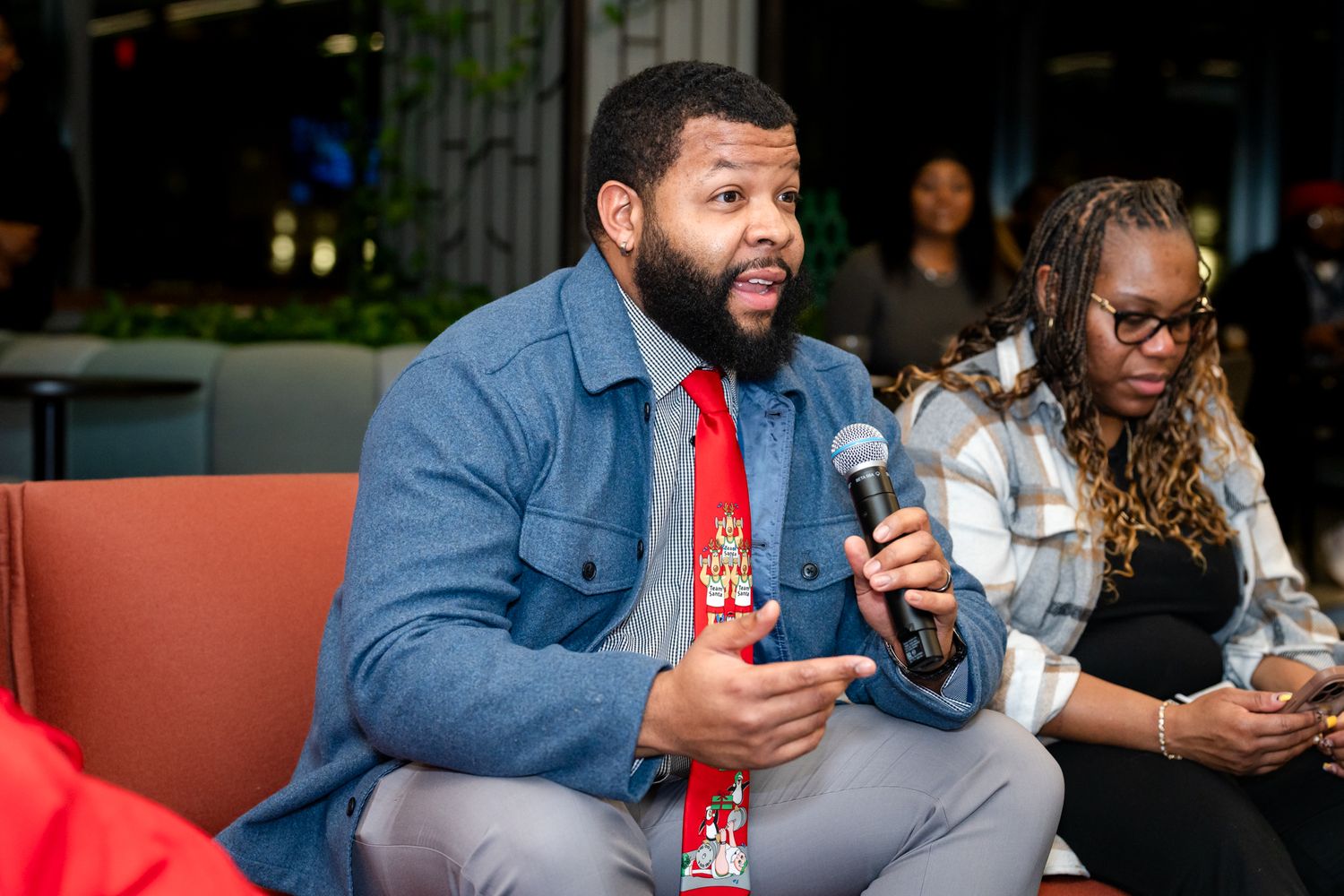 Man speaking into a microphone at a panel discussion event wearing a denim jacket and red tie.