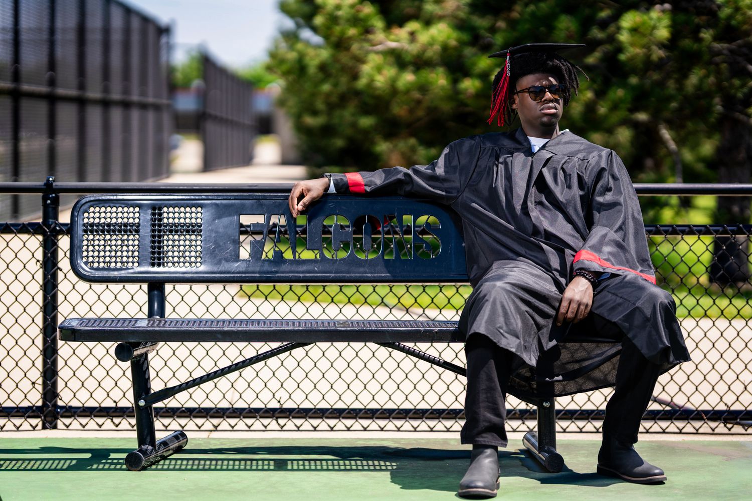 Black male graduate in cap and gown sits on outdoor bench at school campus.