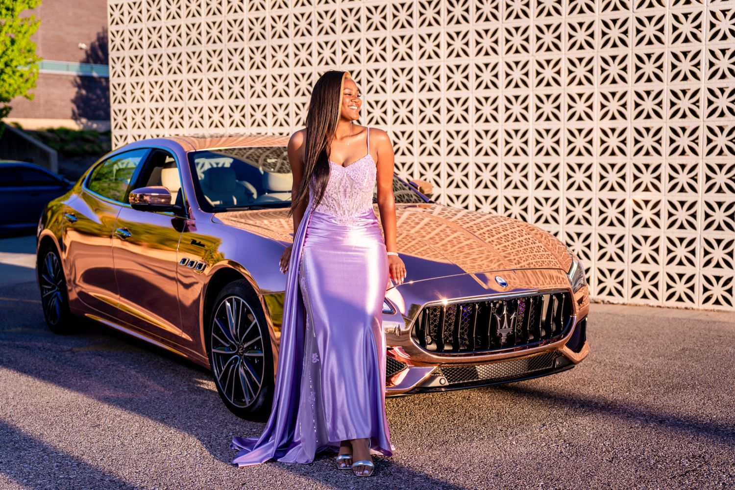 Young woman in lavender prom gown poses beside a gold chrome Maserati luxury vehicle outdoors.