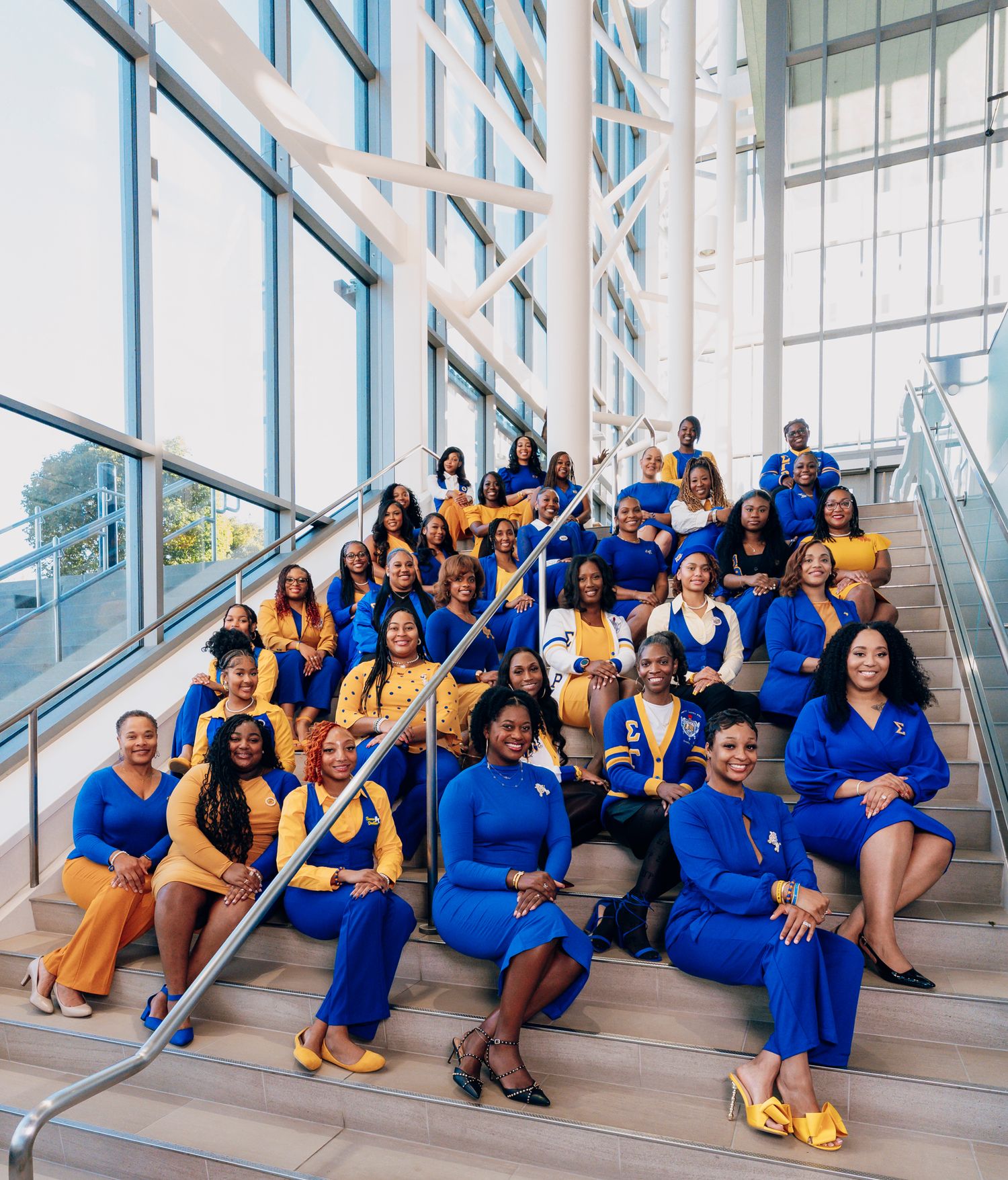 Large group of women in coordinated blue and gold outfits posing together on a modern staircase inside a bright glass-walled building.
