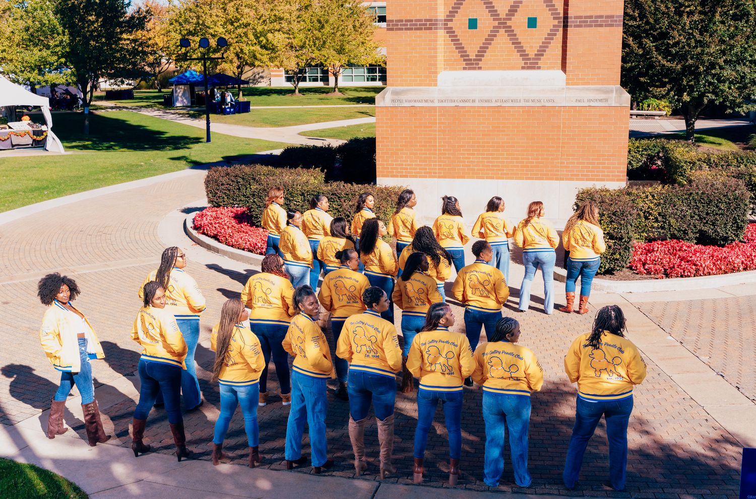 Group of students in matching gold shirts standing in a semicircle on a brick plaza near a campus building with red flower beds.