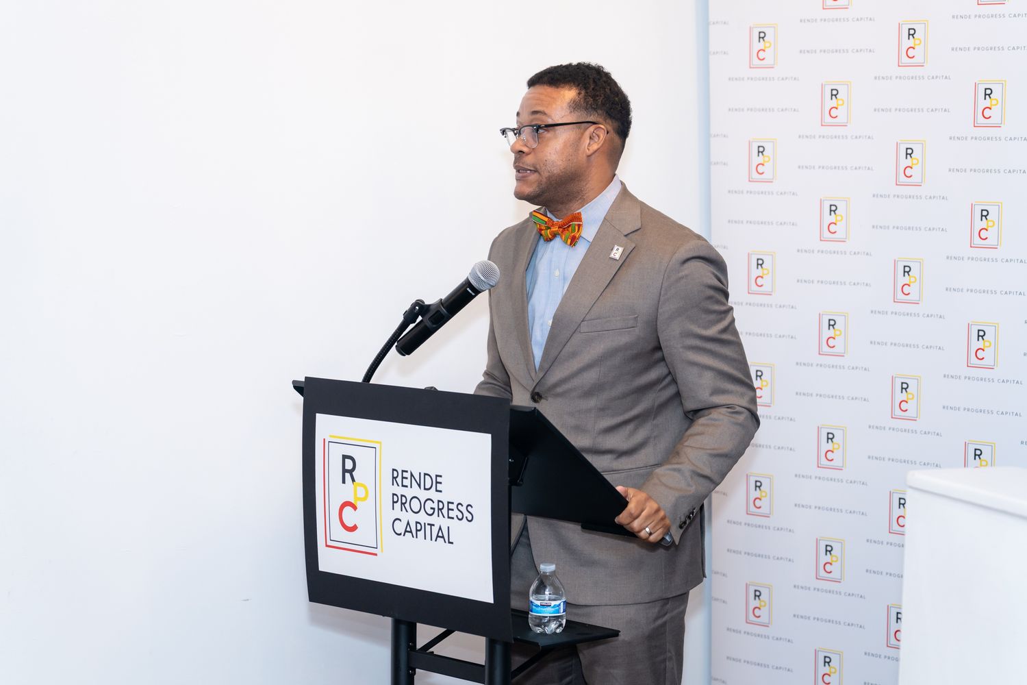 A man in a gray suit and orange bow tie speaks at a Rende Progress Capital podium during an event.