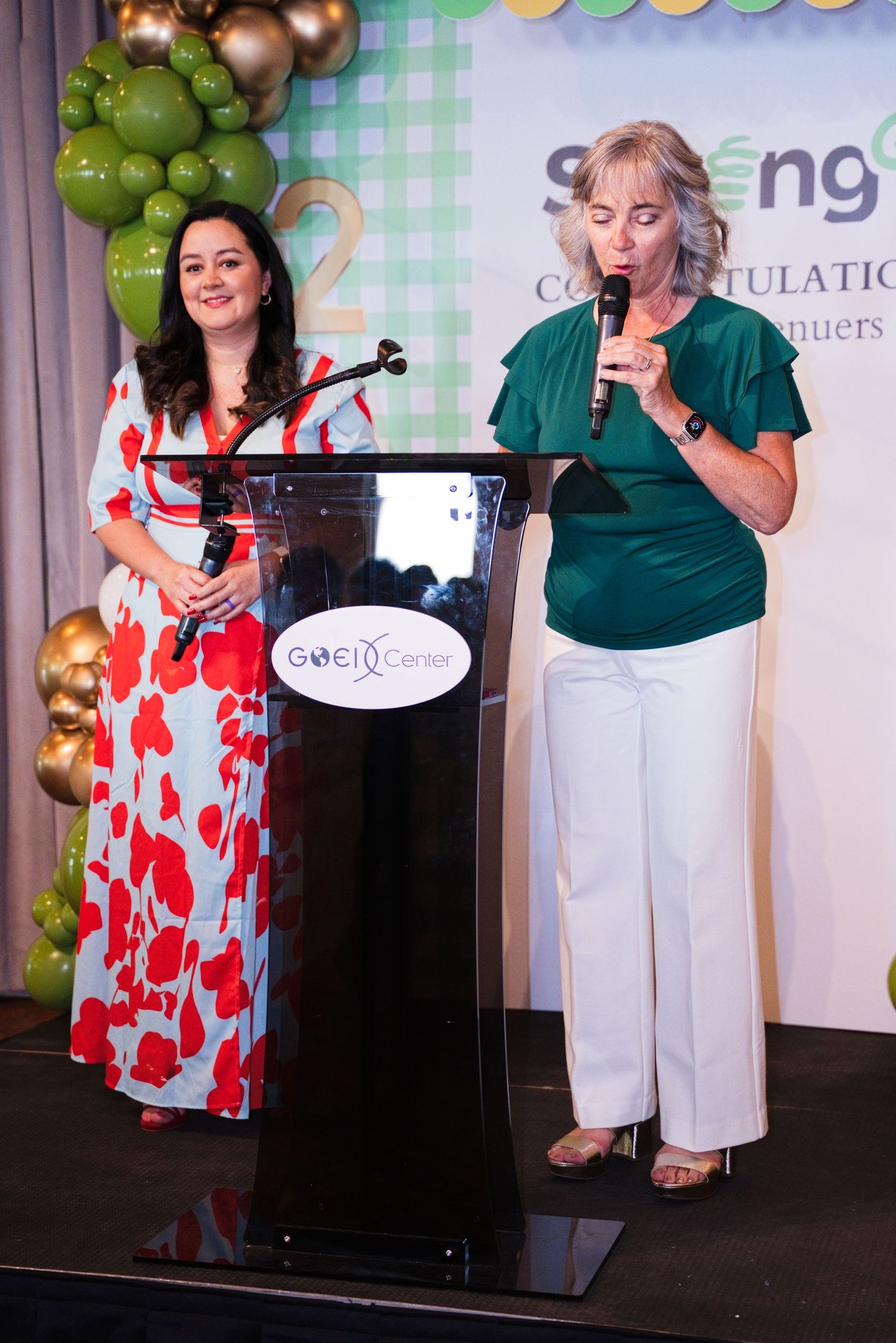 Two women speak at a podium during a congratulatory awards ceremony with green decor.