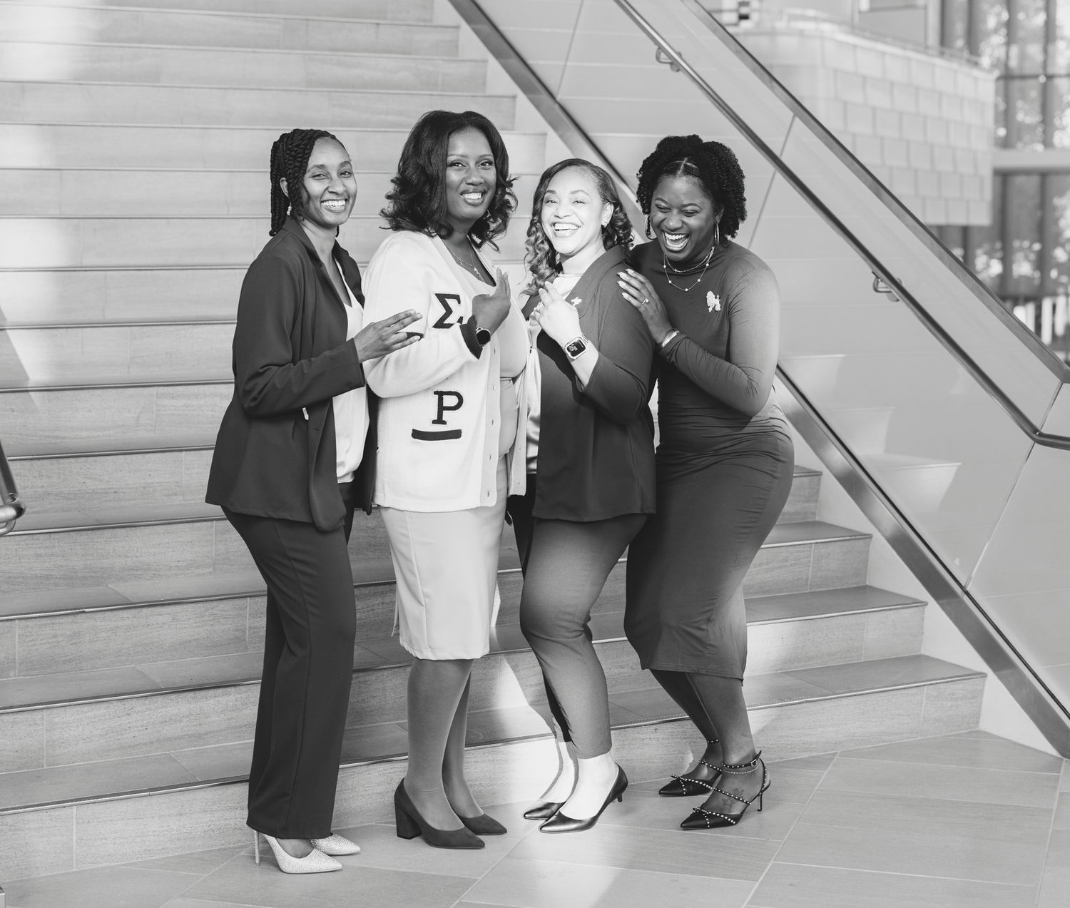 Four smiling women in professional attire pose together on a staircase, one wearing a cream jacket with Greek letters, in black and white.