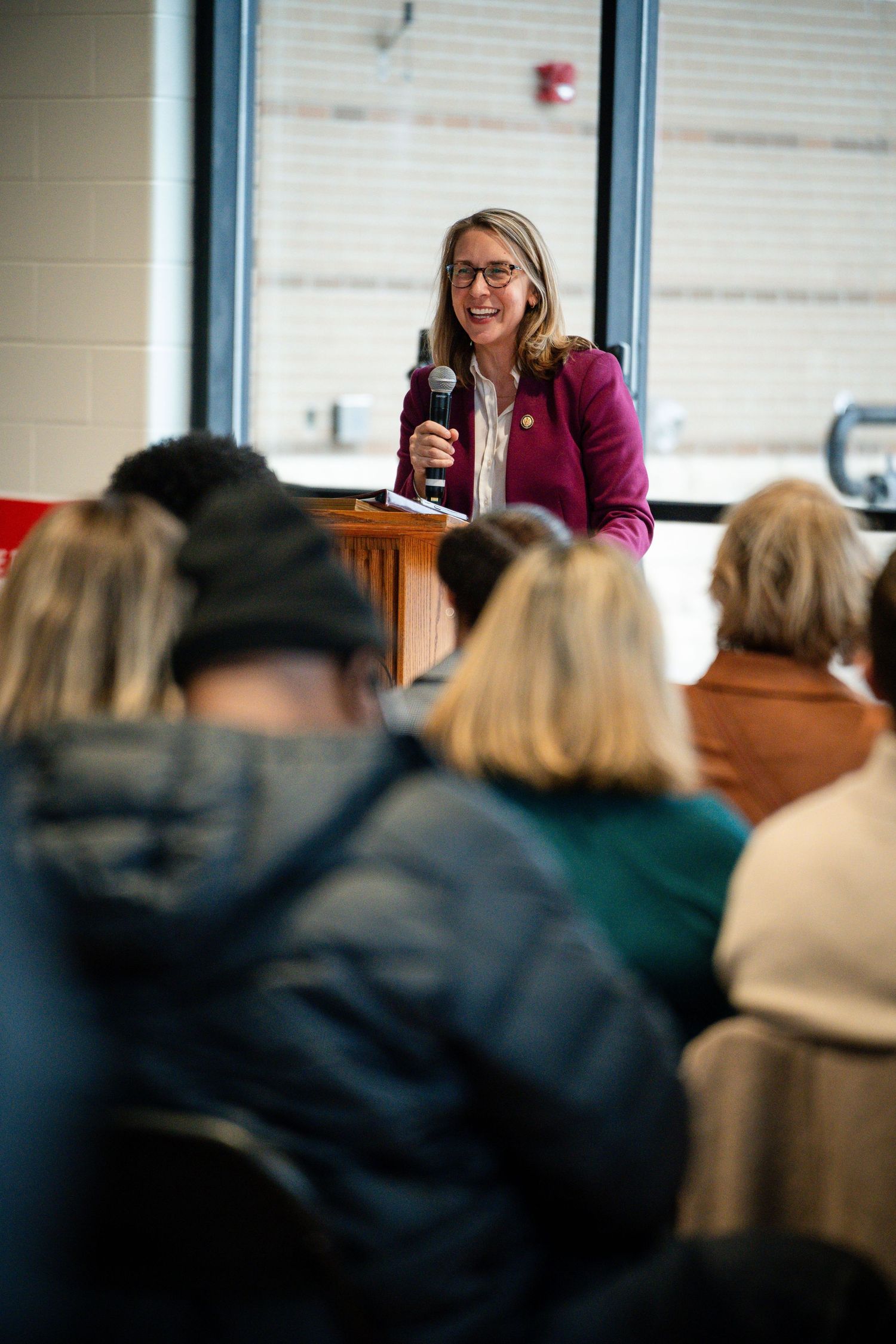 A smiling woman in a purple blazer speaks at a podium with a microphone to a seated audience indoors.