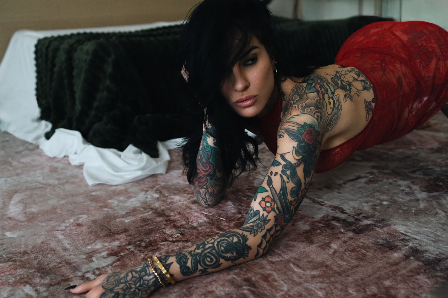 Heavily tattooed woman in red lace lying on a bed, showcasing intricate full-sleeve tattoo artwork.
