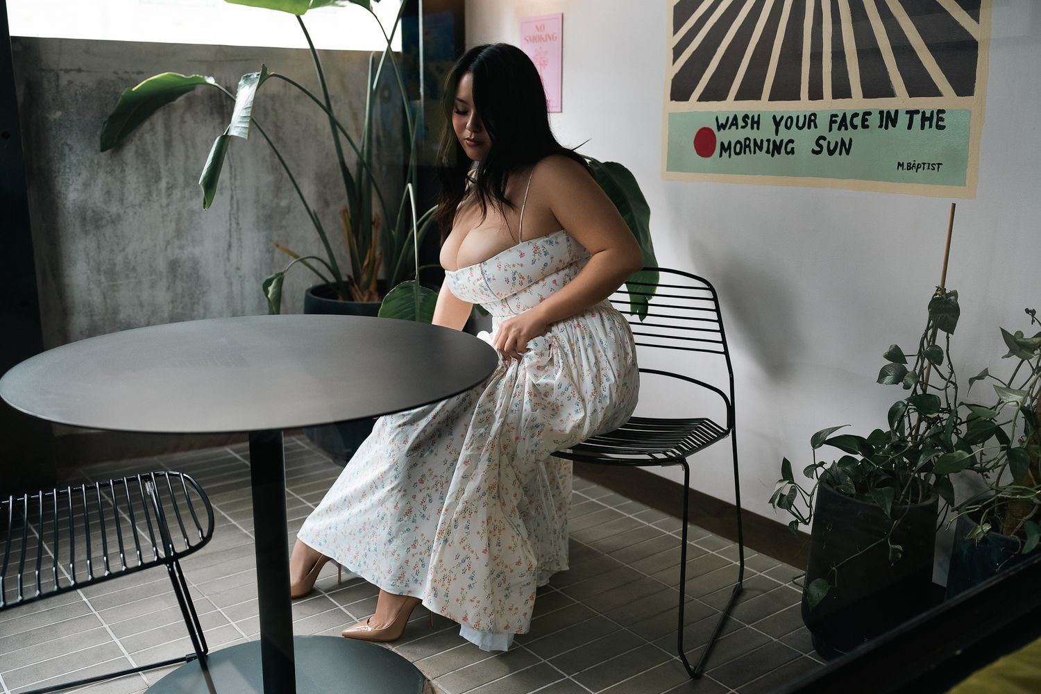 Woman in floral maxi dress sitting barefoot by a round table in a stylish cafe with plants and wall art.