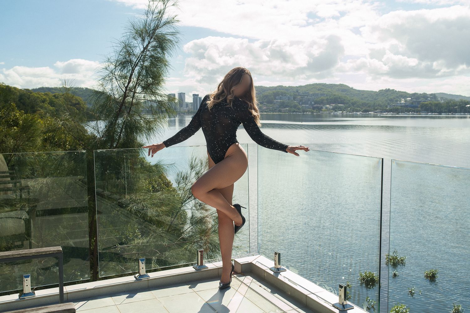Woman in black bodysuit posing on glass balcony railing with scenic lake and hills in background.