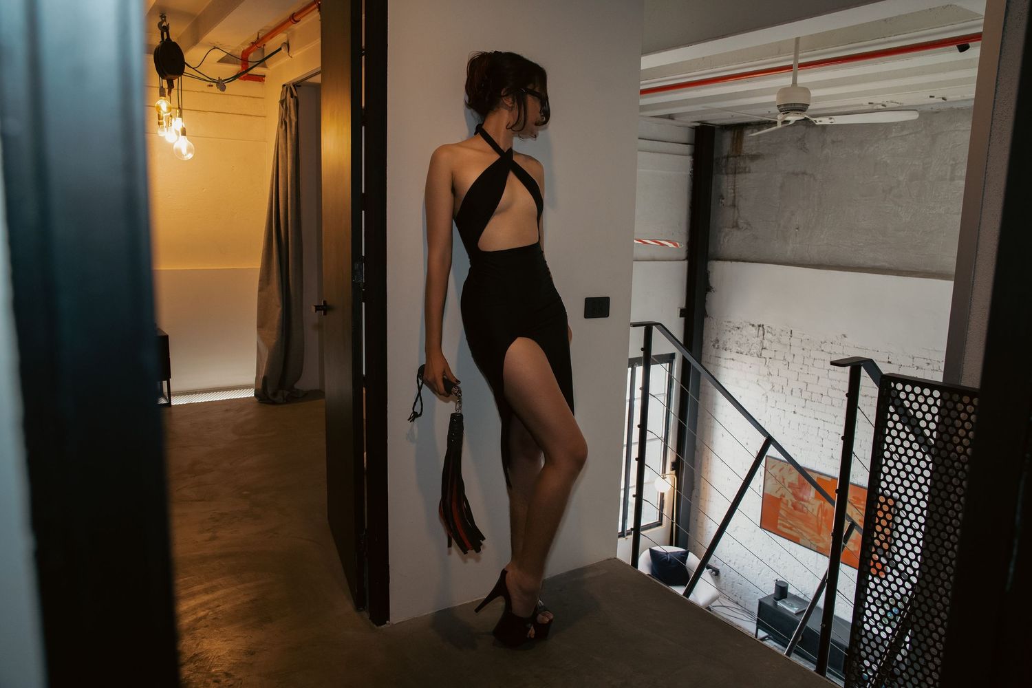 Woman in black backless swimsuit with high slit stands near staircase in industrial loft space.