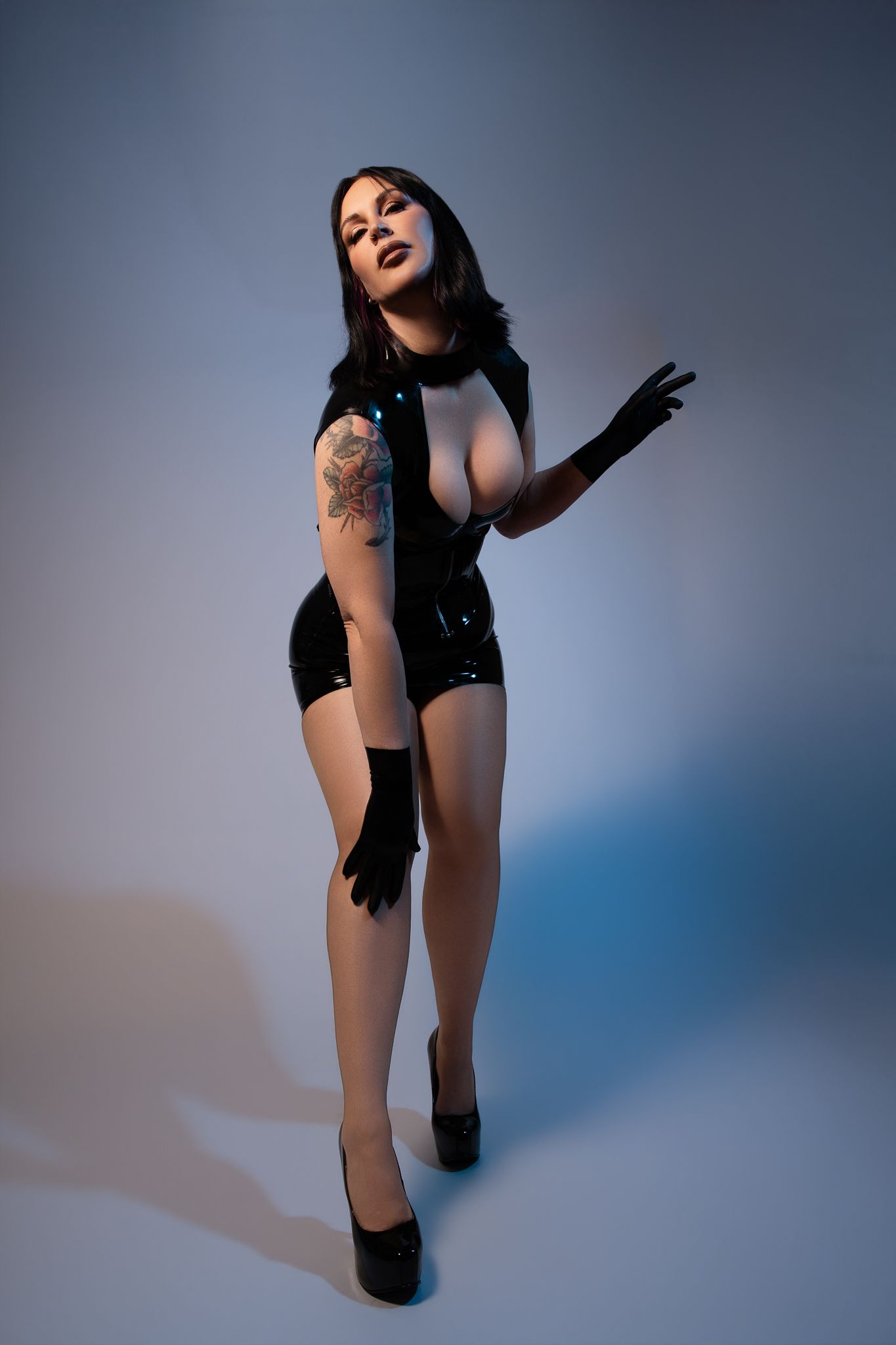 Woman in black latex outfit with gloves and heels posing in studio with blue lighting.