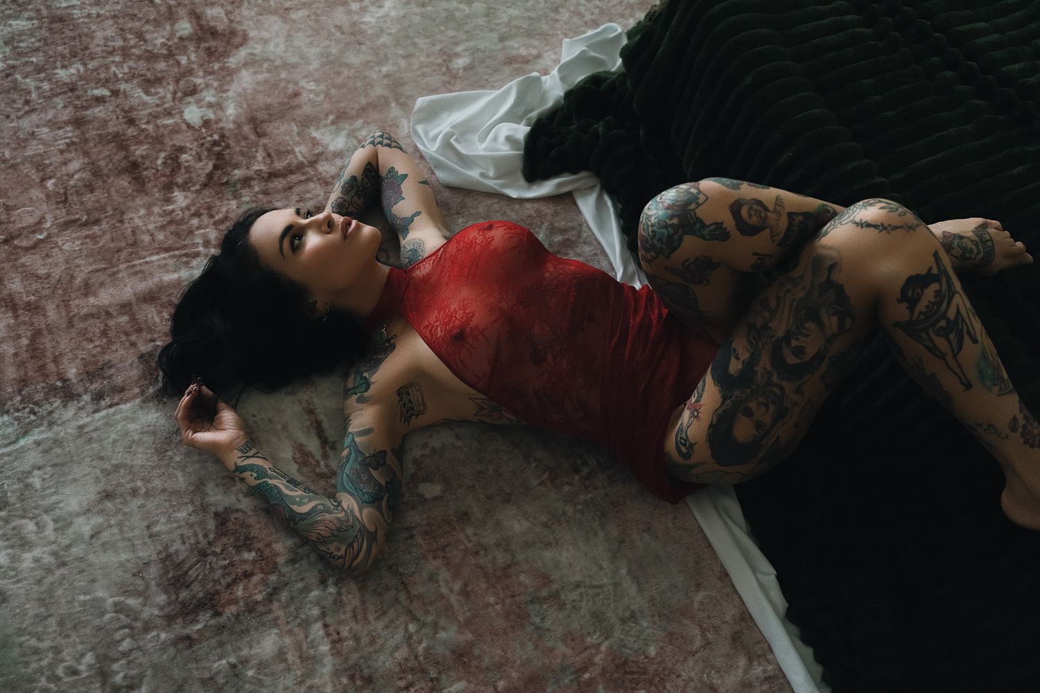 Tattooed woman in red top lying on floor with white fabric, showcasing full sleeve tattoos in moody artistic photo.