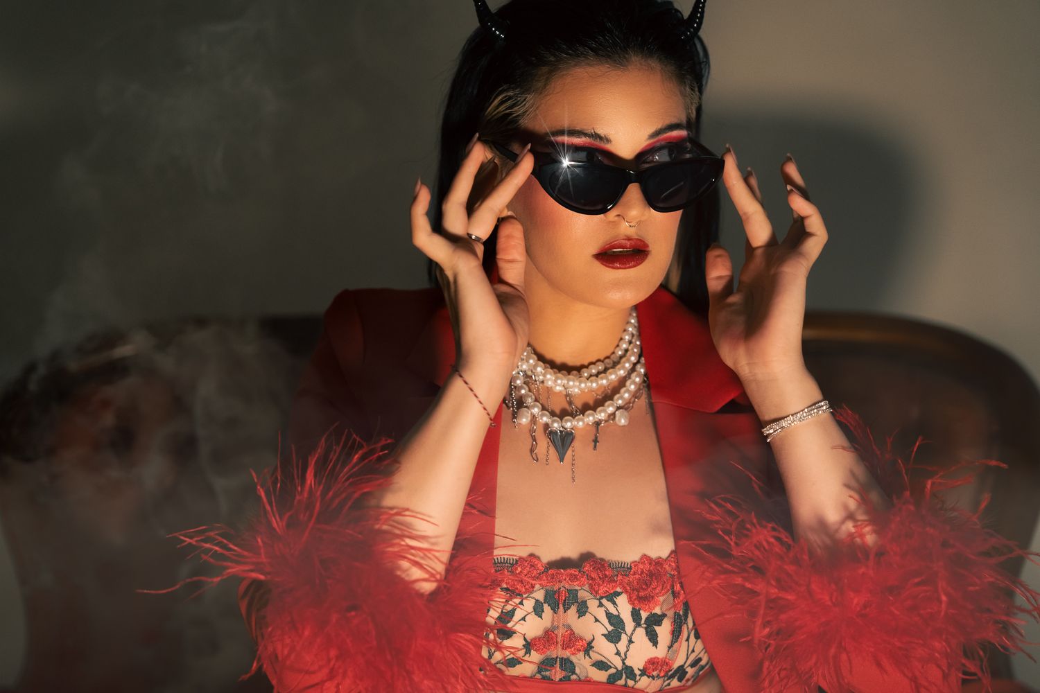 A stylish woman in red feather jacket adjusts cat-eye sunglasses, wearing layered necklaces and devil horns.