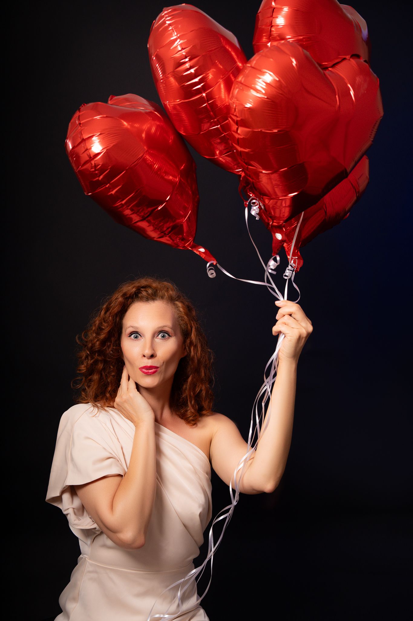 Woman in white dress poses playfully with red heart-shaped balloons against dark background.