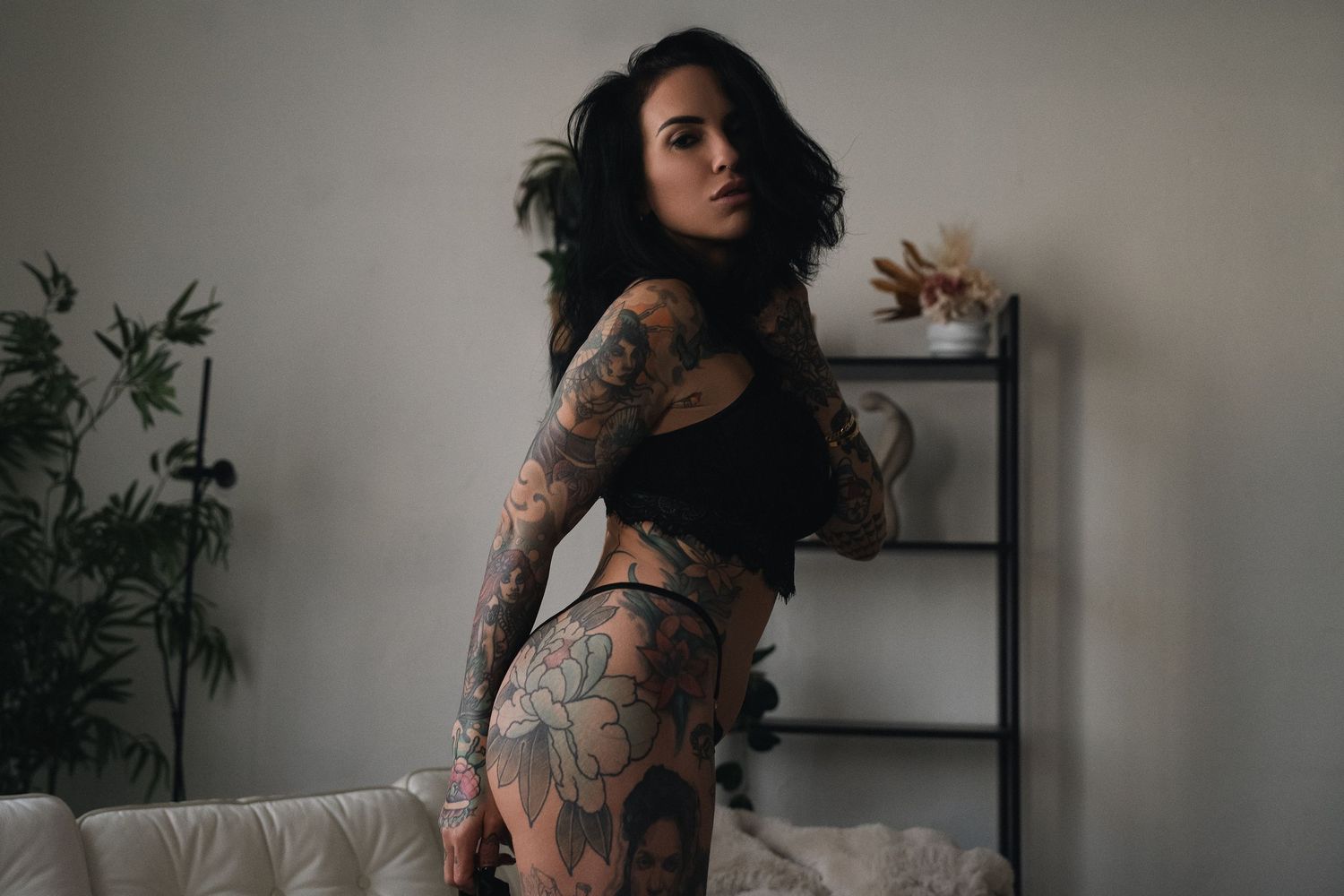 Tattooed woman in black crop top and underwear posing in a modern room with plants and shelving.