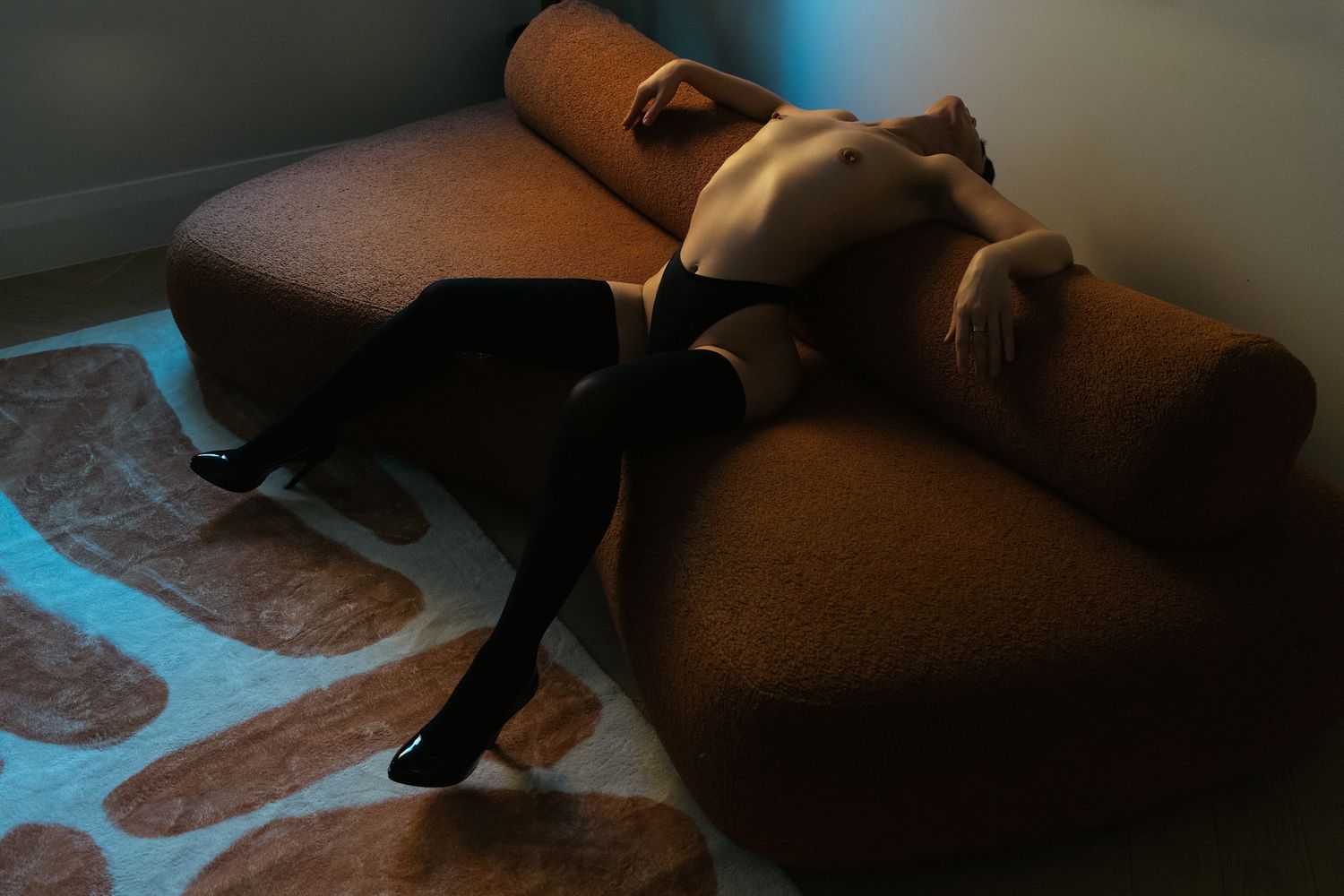 Person in black stockings and heels reclining on a brown curved sofa on a patterned rug.