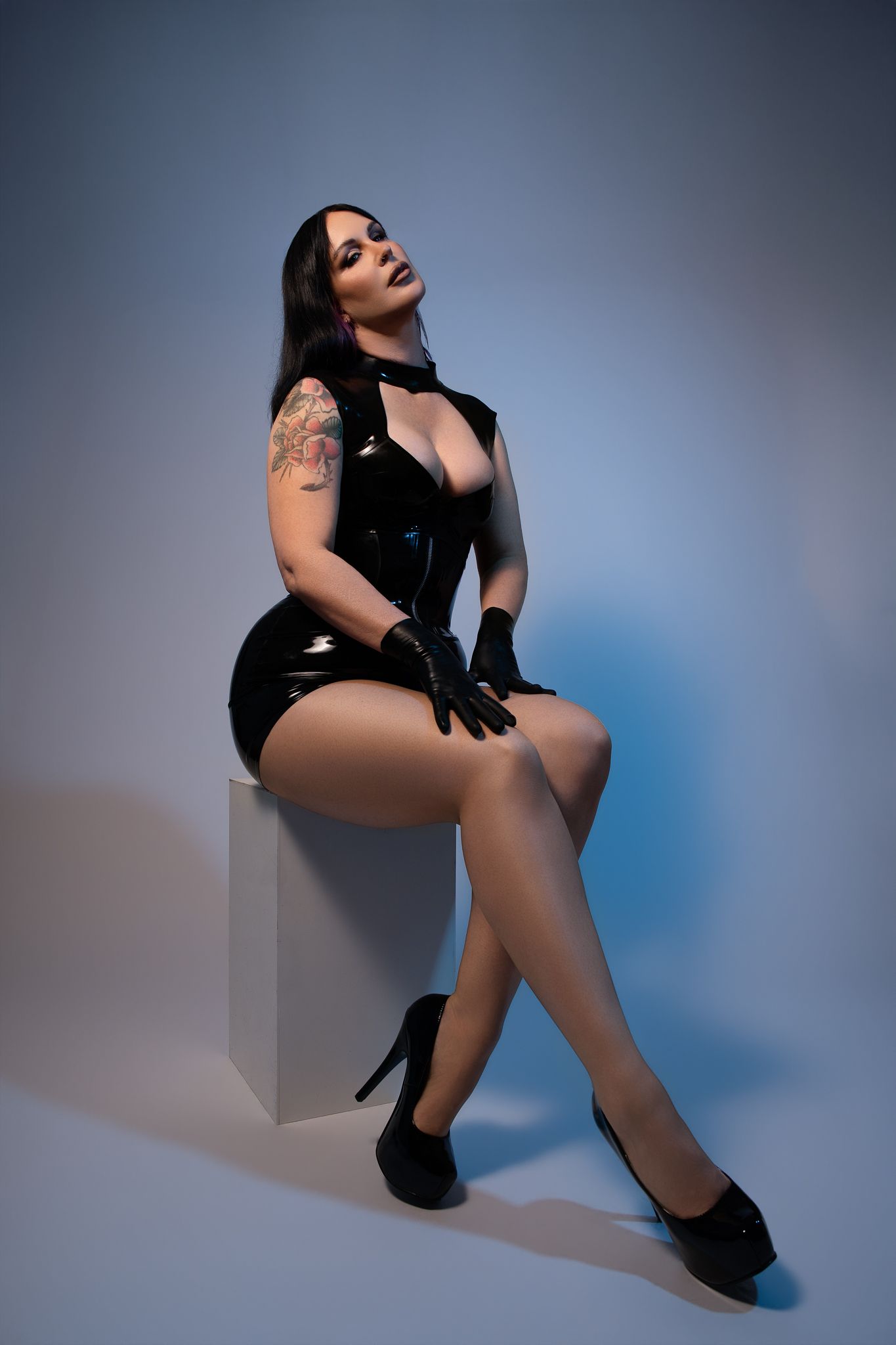 Woman in black latex dress and gloves posing seated on white cube in studio with blue background.