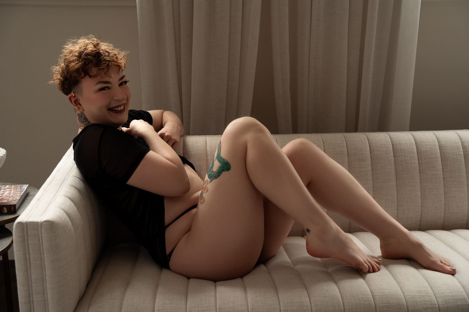 Young woman with curly hair smiling while relaxing on a white sofa in a dimly lit room.