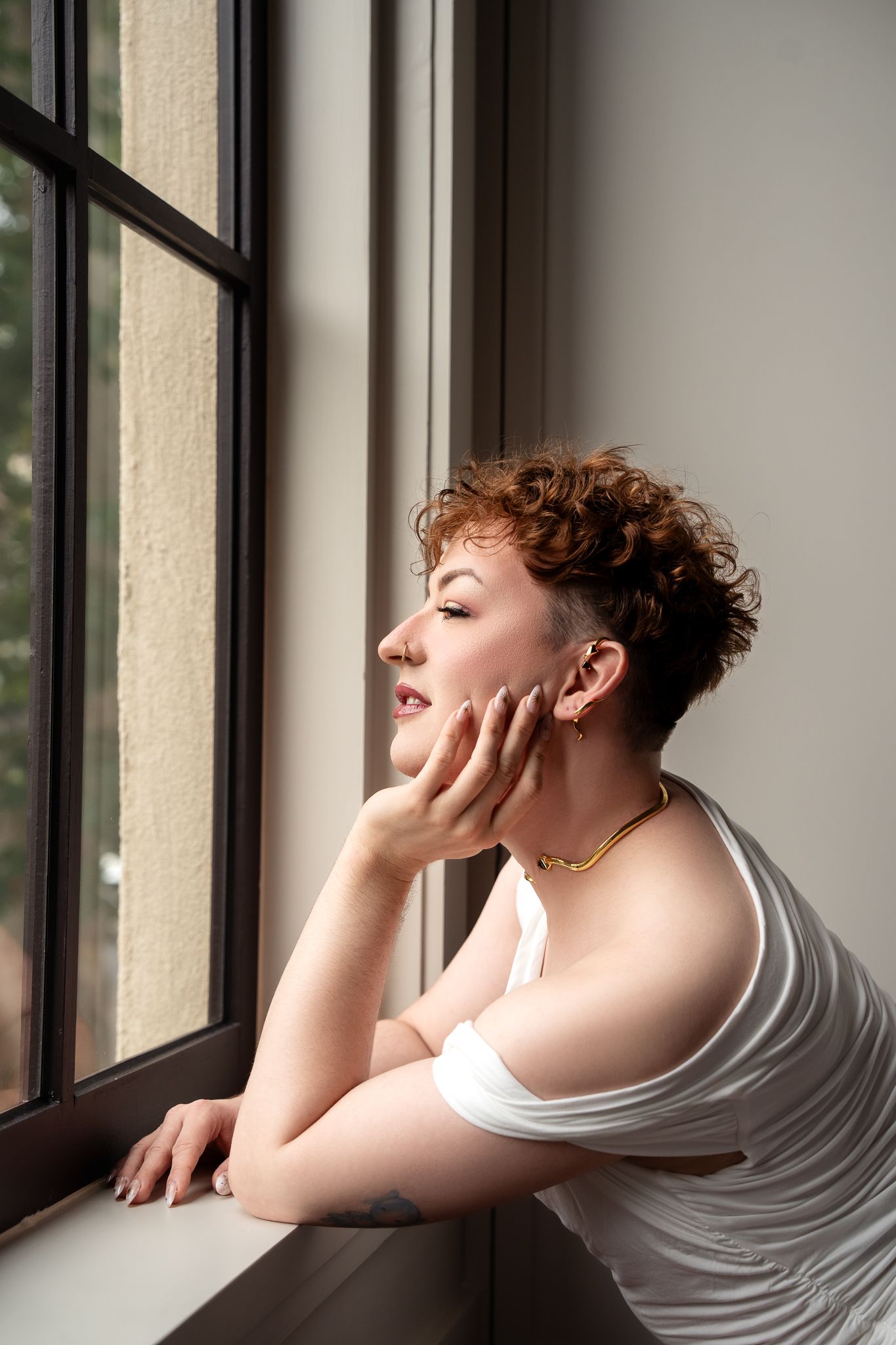 Young woman with curly red pixie cut leans on windowsill, wearing white top and gold necklace, gazing outside thoughtfully.