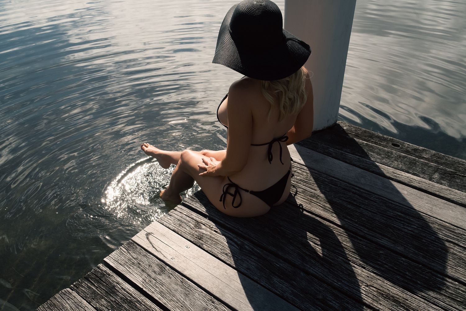 Woman in black bikini and sun hat sitting on wooden dock, splashing feet in calm water on sunny day.