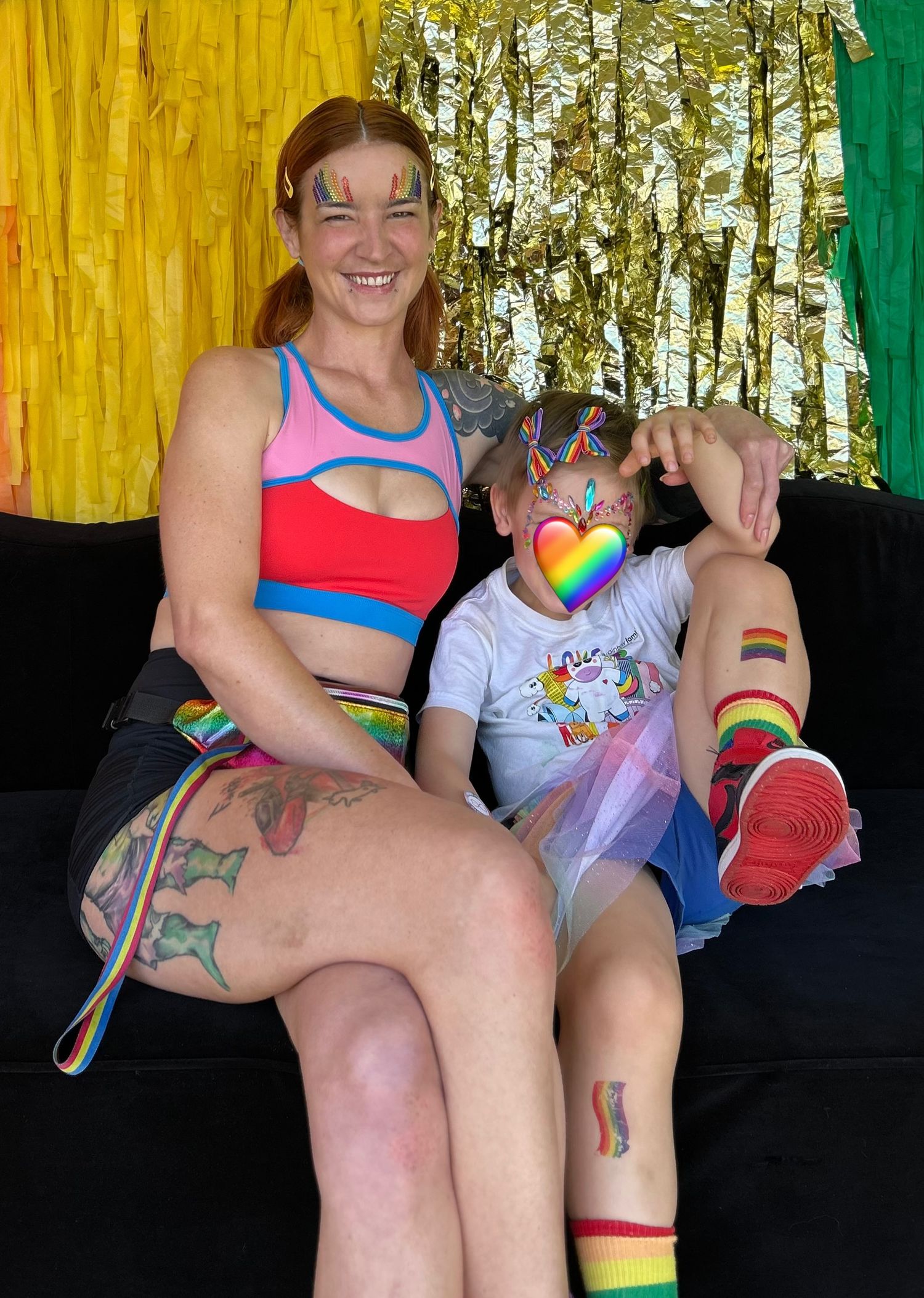 Two people wearing rainbow-themed pride attire and face paint sit together on a couch against a colorful backdrop.