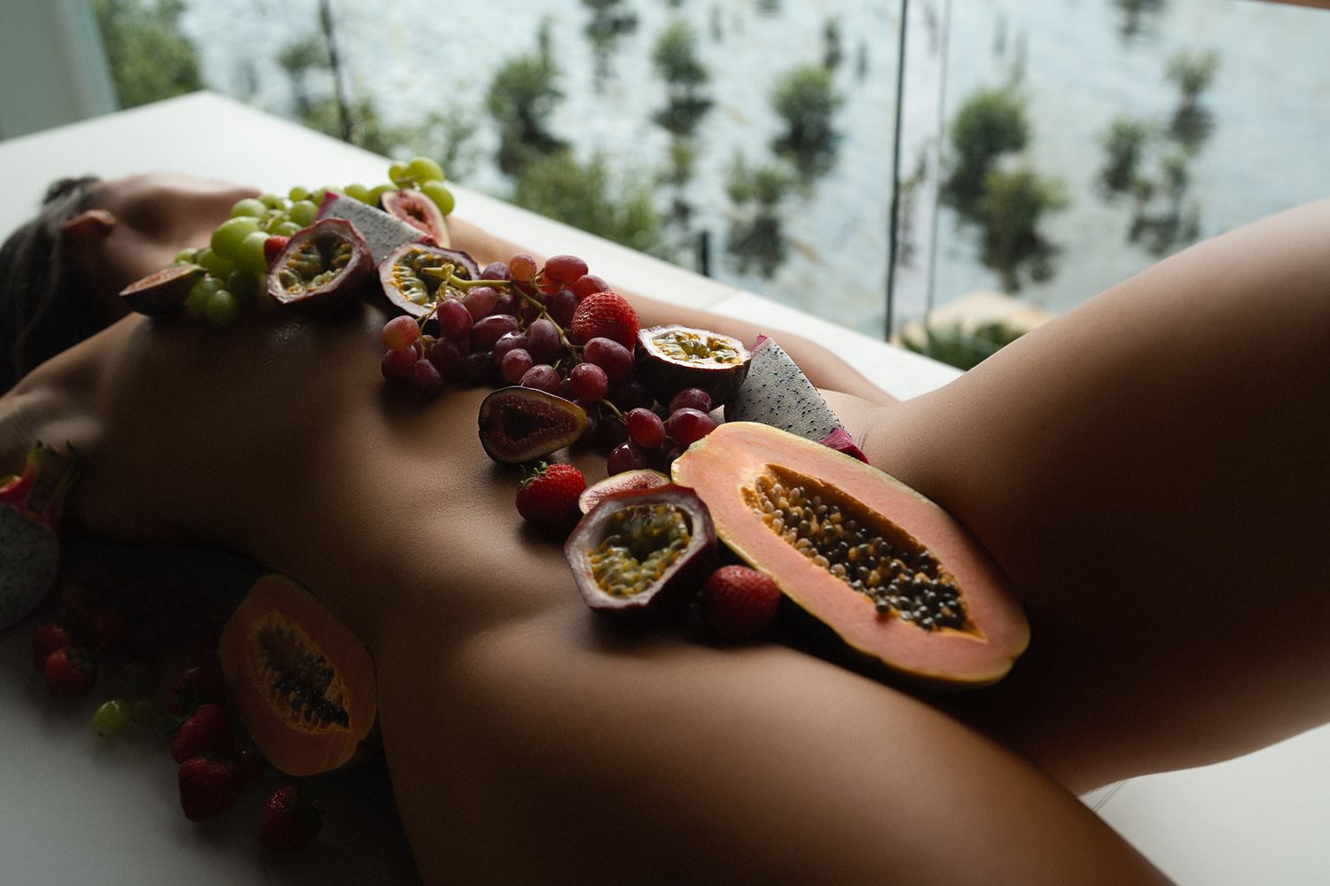 Tropical fruits including papaya, grapes, and passion fruit artfully arranged on a white surface near a window.