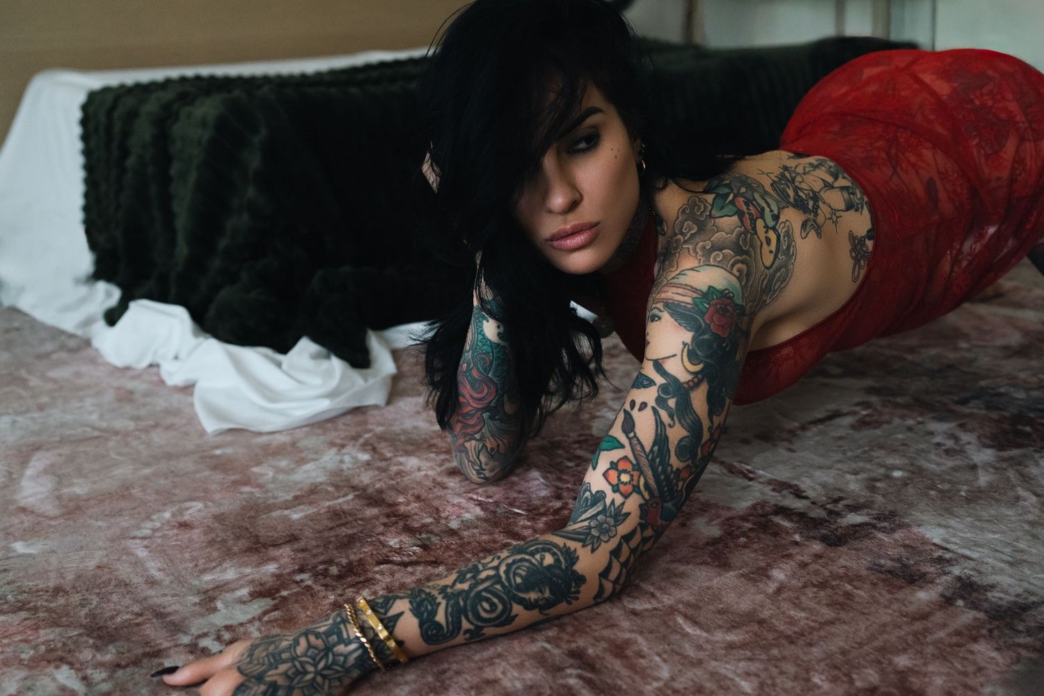 Heavily tattooed woman in red lace lying on a bed, showcasing intricate full-sleeve tattoo artwork.