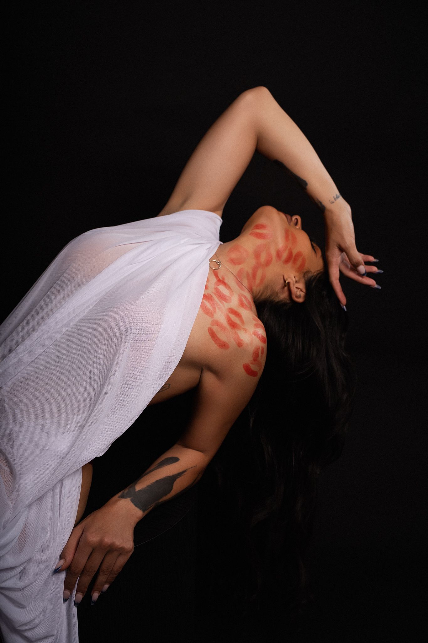 Woman in white draped dress with red lipstick kiss marks on chest, posing dramatically against black background.