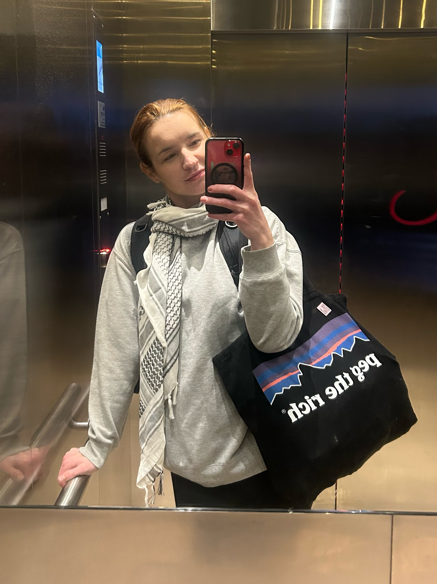 Person wearing light grey hoodie takes elevator mirror selfie while holding black shopping bag with minimal design.
