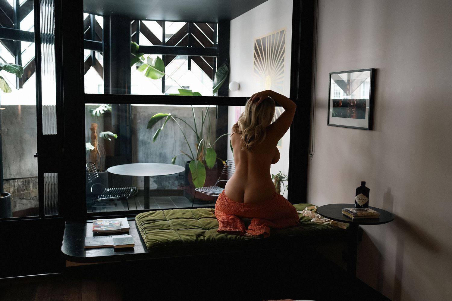 Woman sitting on a bed facing large windows with green plants and outdoor patio view in a modern bedroom.
