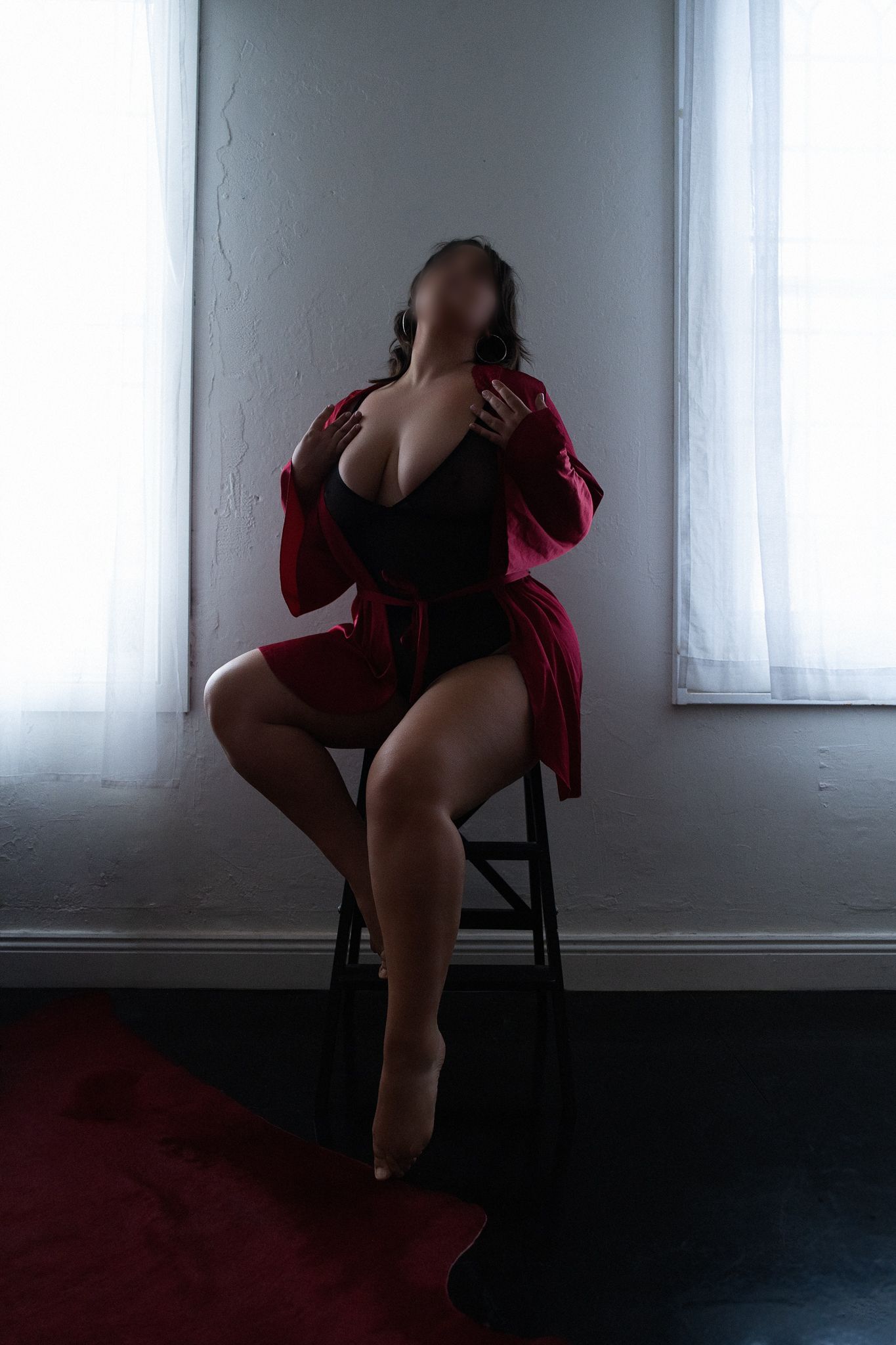 A woman in a black bodysuit and red robe sits on a stool by a bright window in a moody boudoir photo.