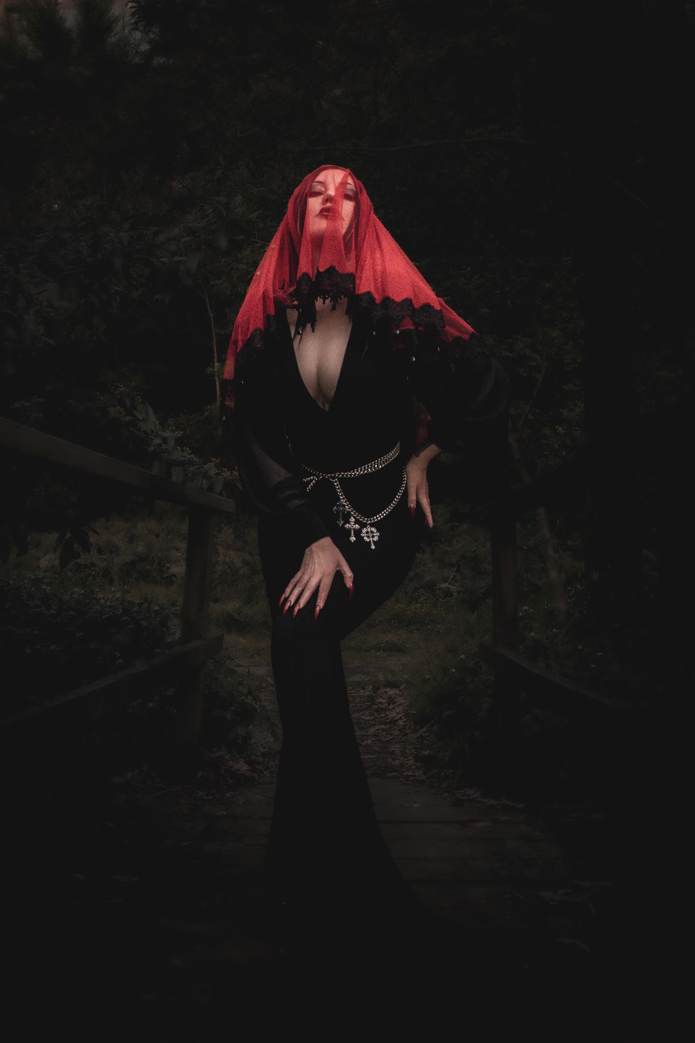 Figure in a flowing black dress and red headpiece poses dramatically against a dark forest background in a gothic style.